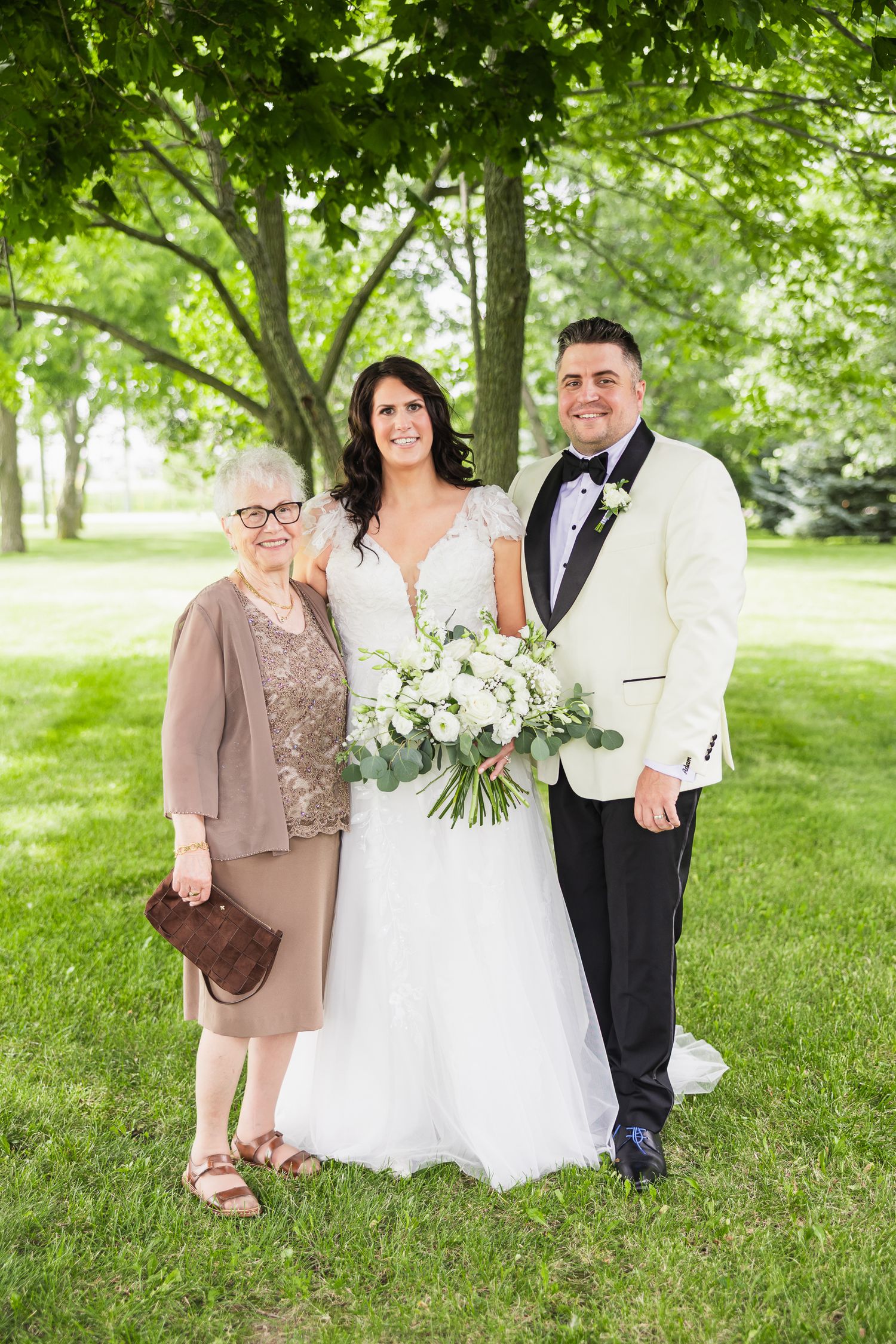 Bride in white gown holds bouquet with groom in ivory tuxedo and elderly woman in green park setting.