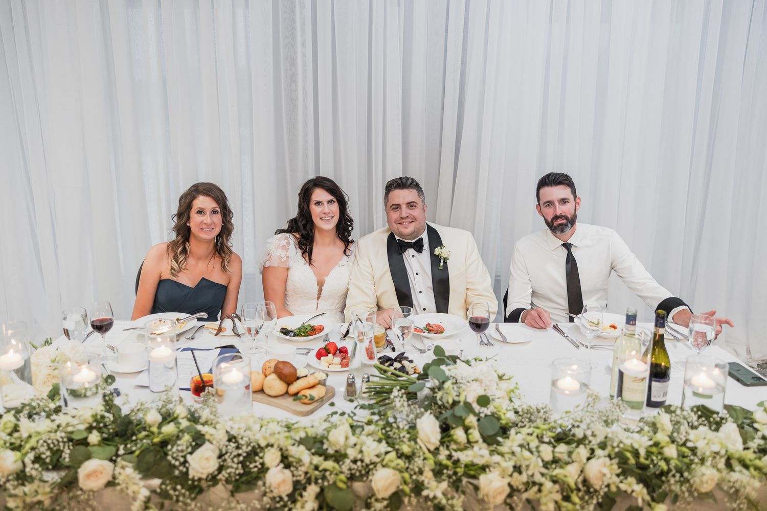 Four wedding party members smile at elegantly decorated head table adorned with white floral centerpieces.