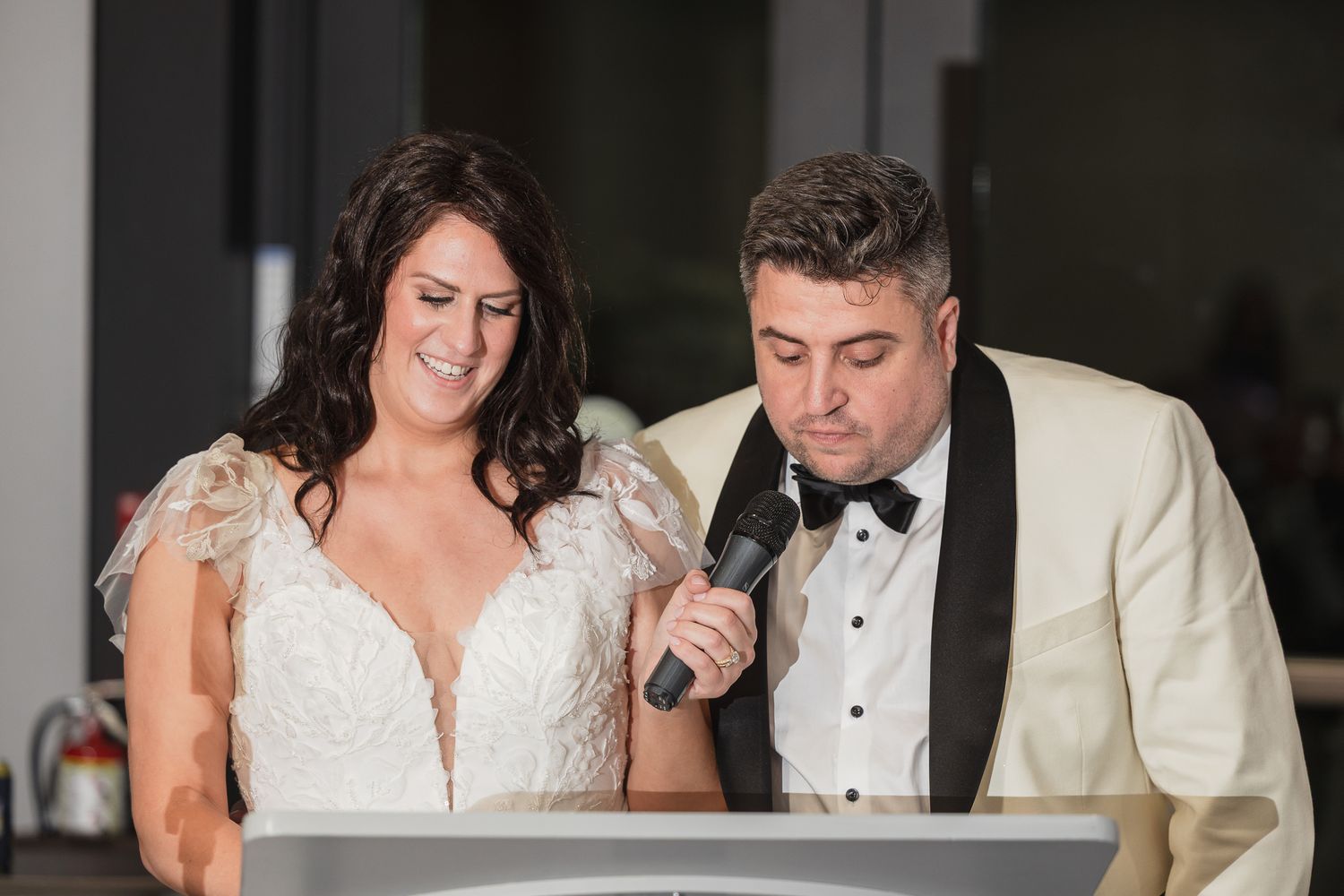 Bride and groom sharing a microphone at wedding reception podium, wearing white dress and ivory tuxedo.