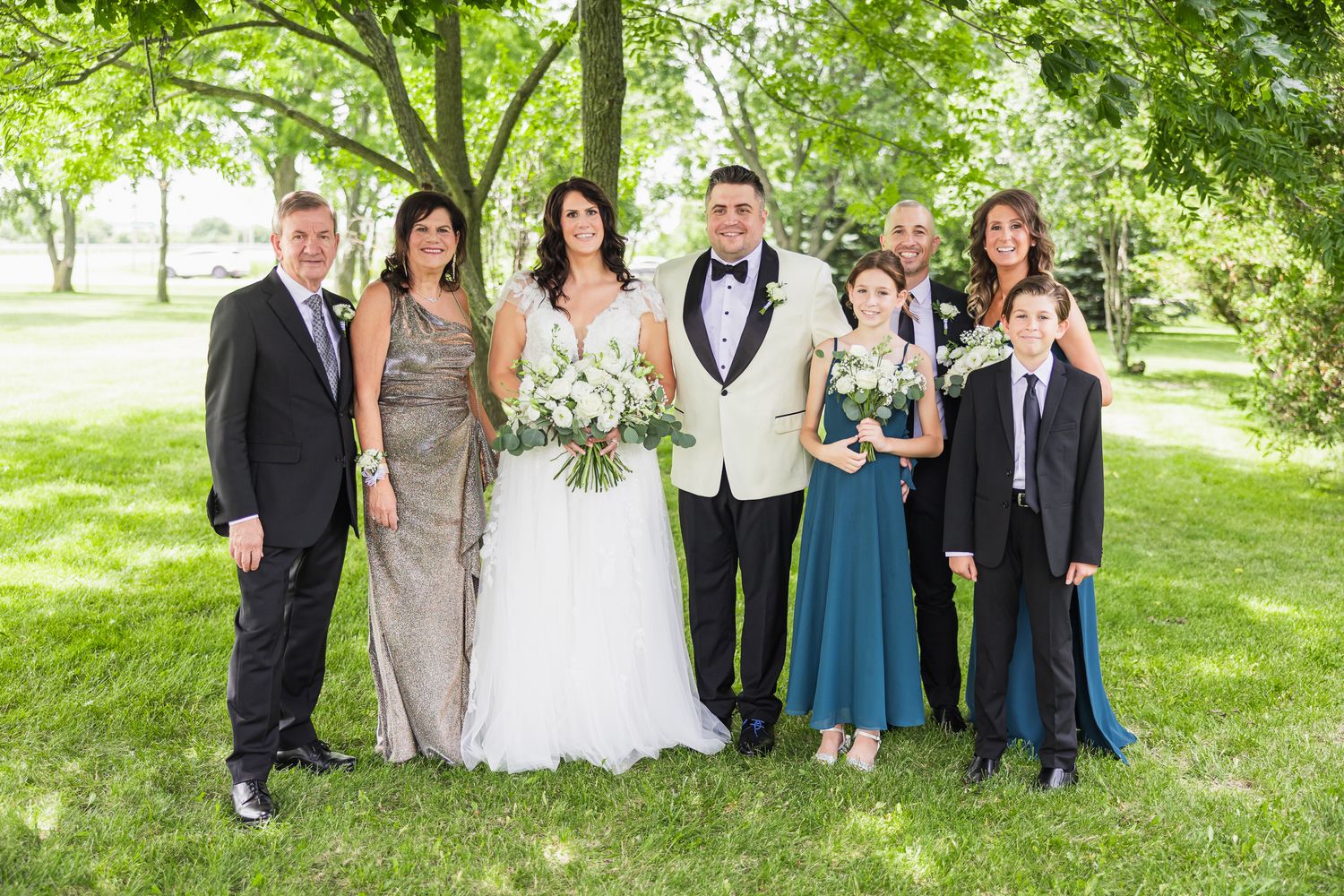 Wedding family portrait on green lawn with bride in white gown and groom in ivory tuxedo jacket.