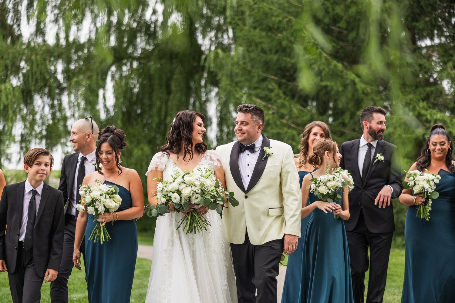 Wedding party walking outdoors, bridesmaids in teal dresses, bride in white gown, groom in ivory tuxedo.