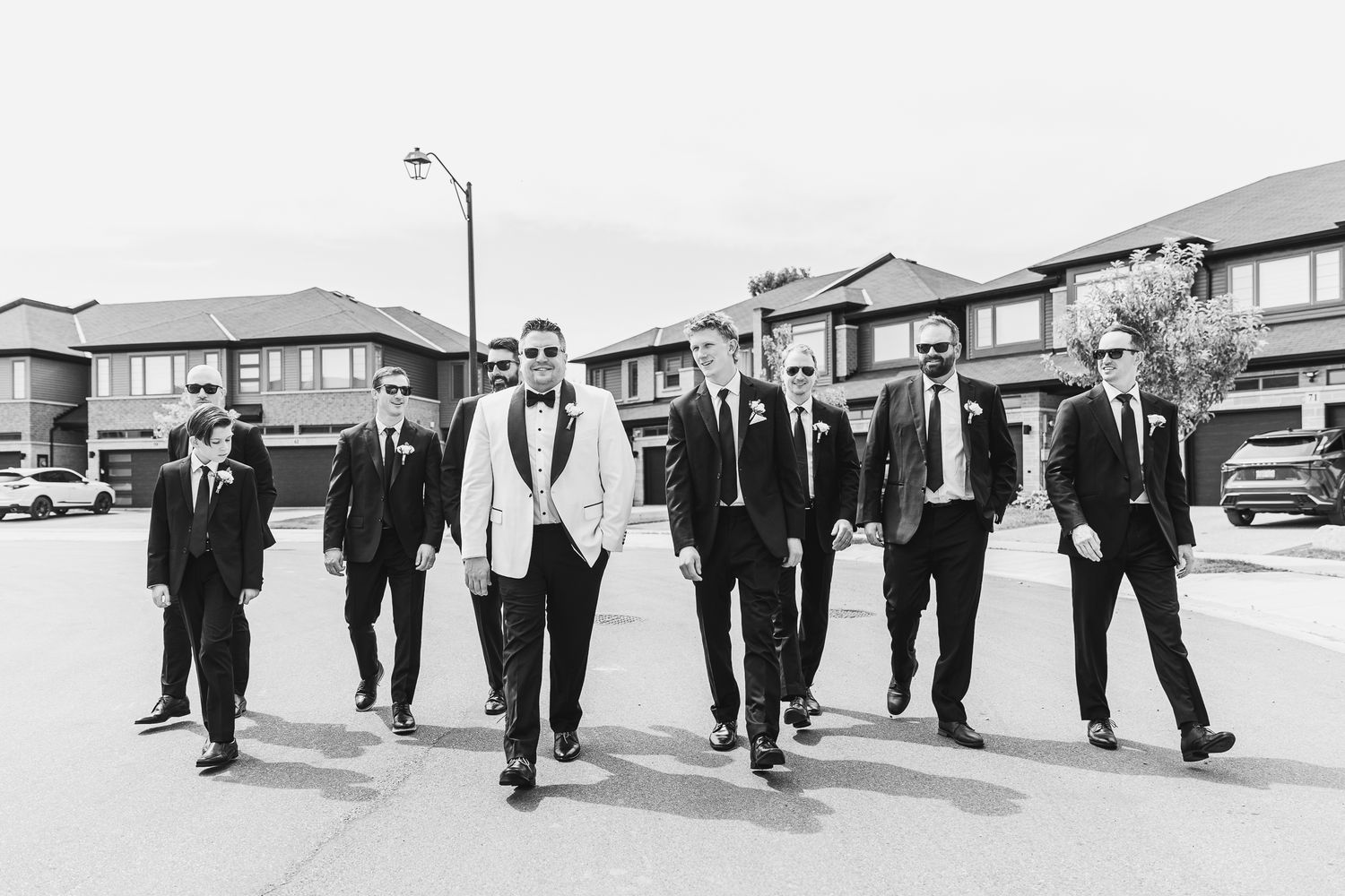Groom in white tuxedo jacket walks with groomsmen in black suits down a suburban street in black and white photo.