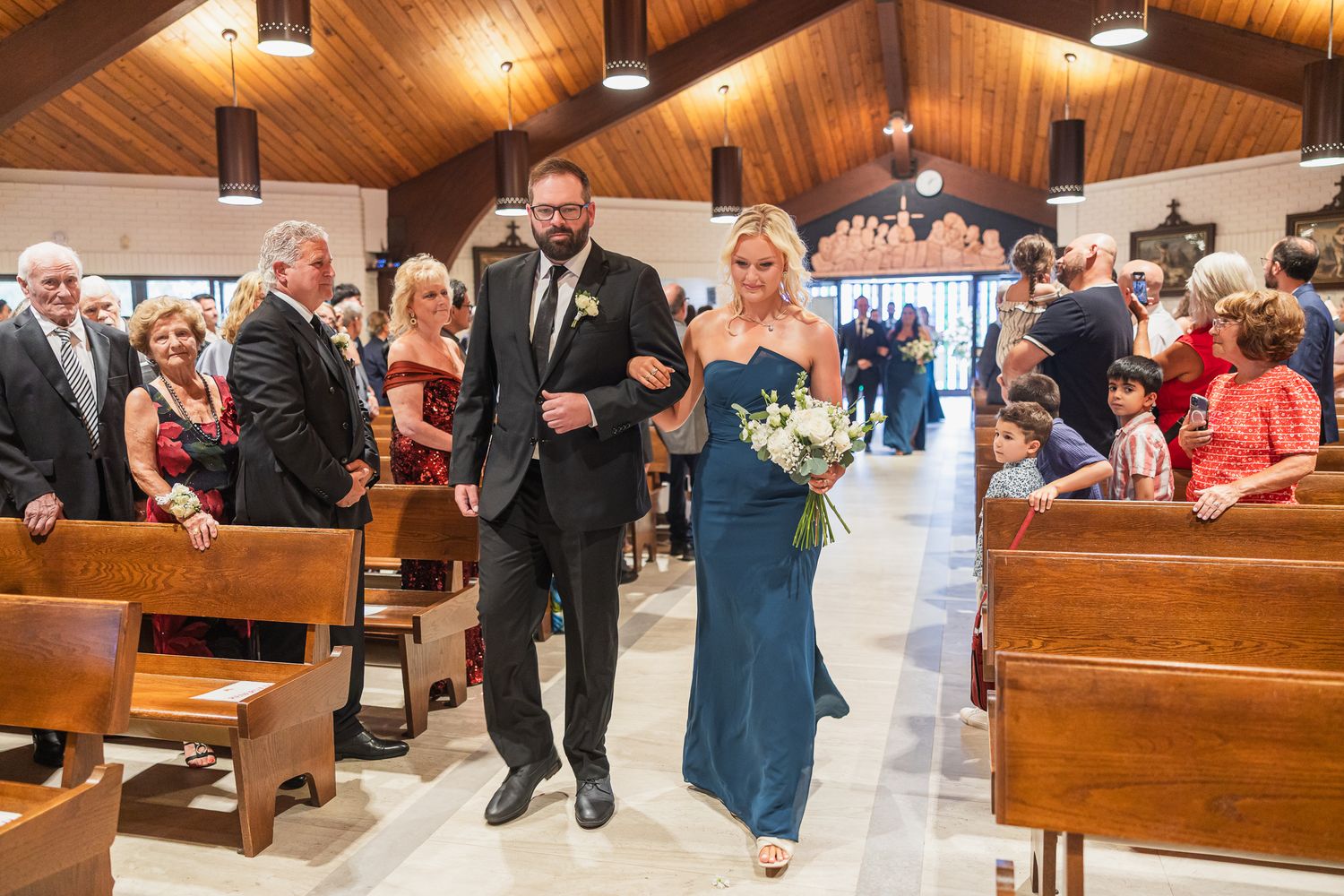 Groomsman escorts a bridesmaid in a teal gown down the church aisle during a wedding ceremony.