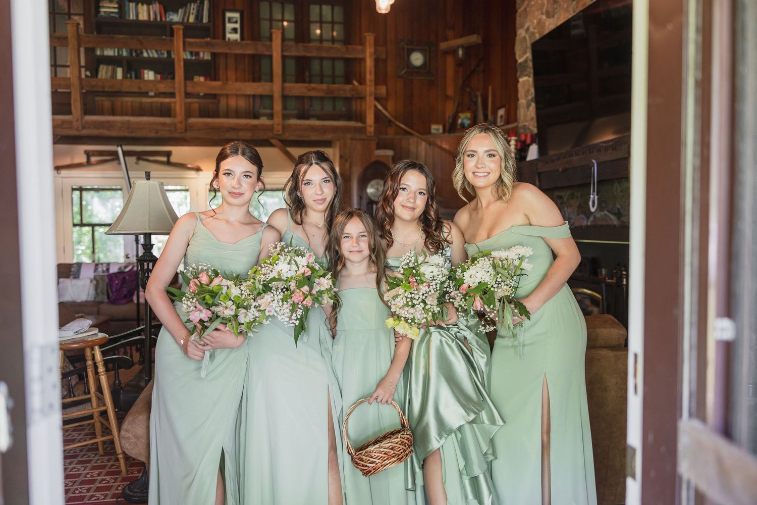 Four bridesmaids and flower girl in sage green dresses holding bouquets in rustic barn venue.