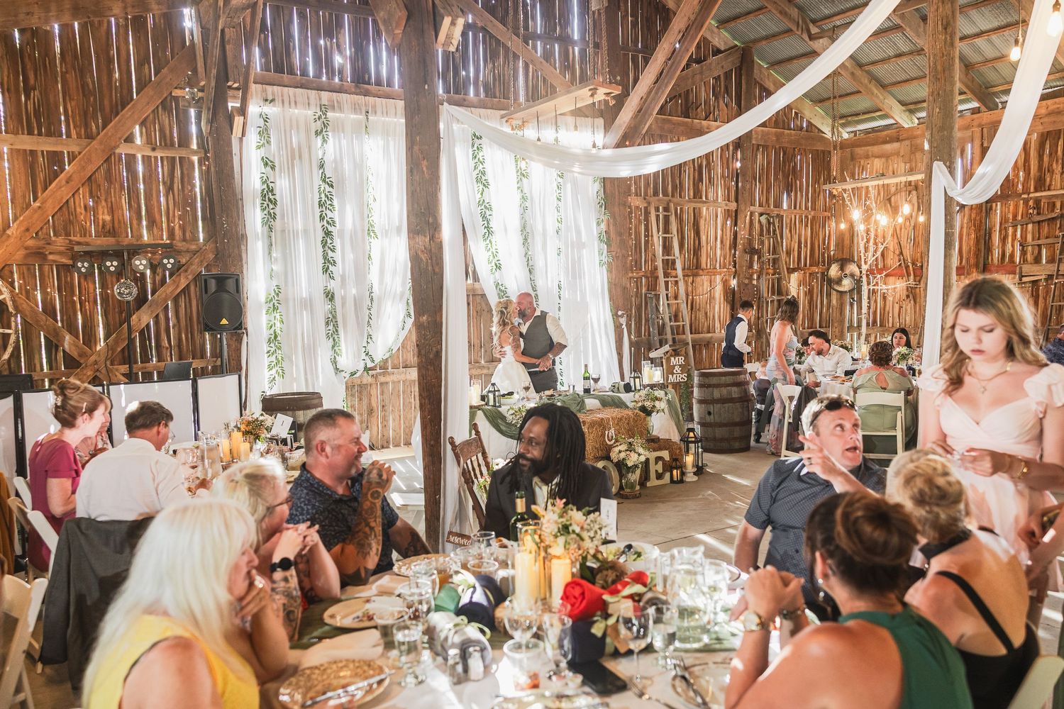 Guests celebrate at a rustic barn wedding reception with draped white fabric, floral centerpieces, and warm ambient lighting.