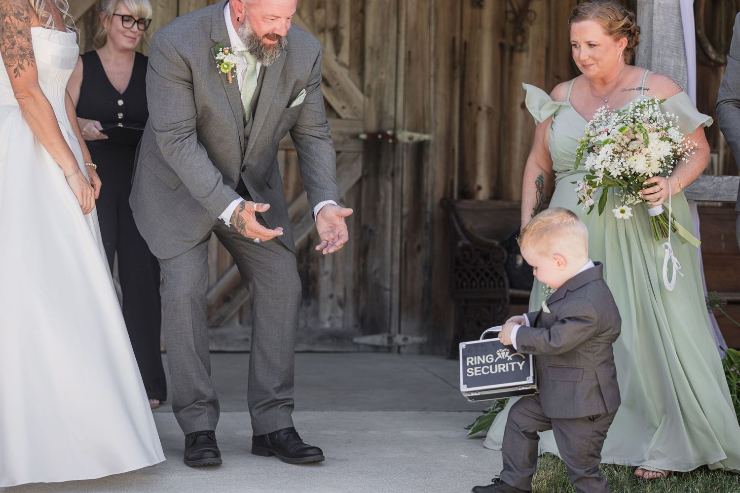 Cute toddler ring bearer carrying sign approaches groom at rustic barn wedding ceremony entrance.