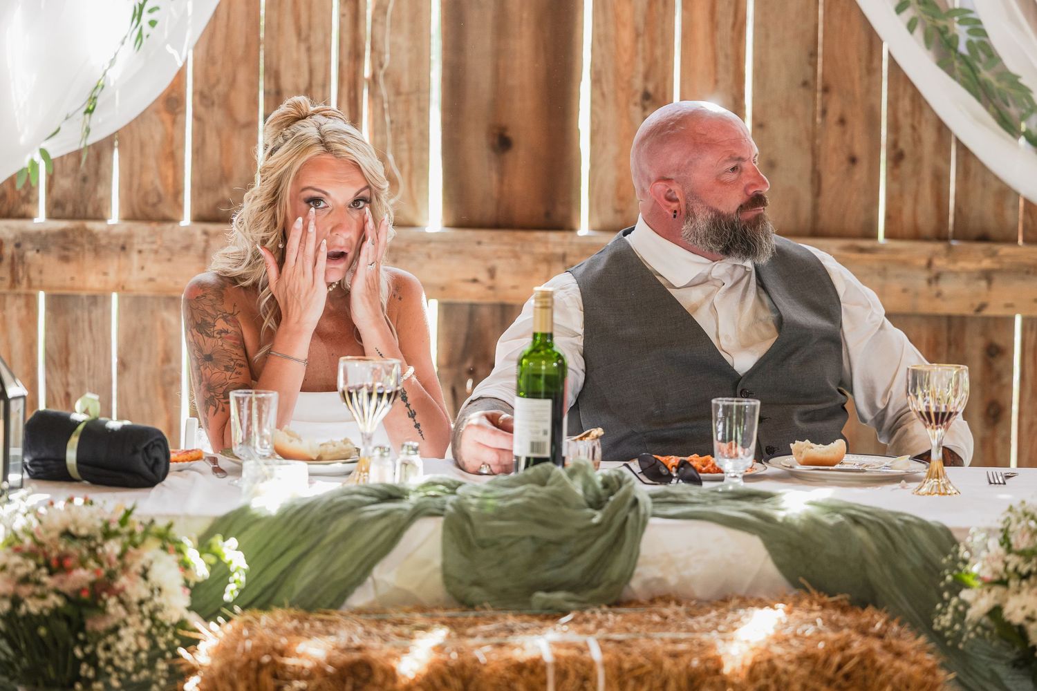 Bride with hands on face in shock sits beside groom at rustic barn wedding reception head table.
