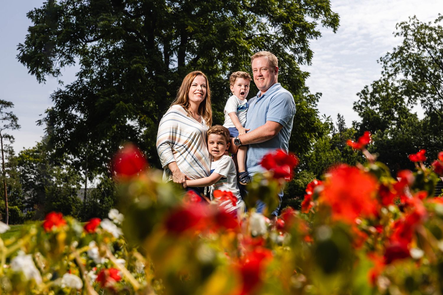 A family poses together in a garden with vibrant red flowers blooming in the foreground and a large tree in the background.