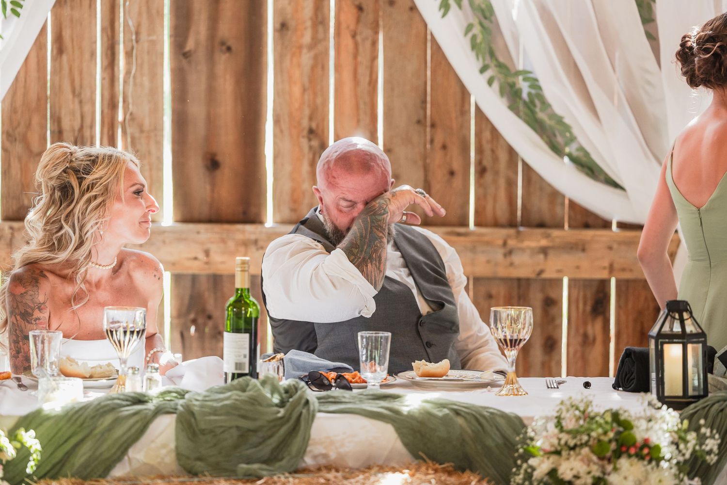 Emotional wedding reception moment with groom embracing guest at rustic barn venue with elegant table settings.