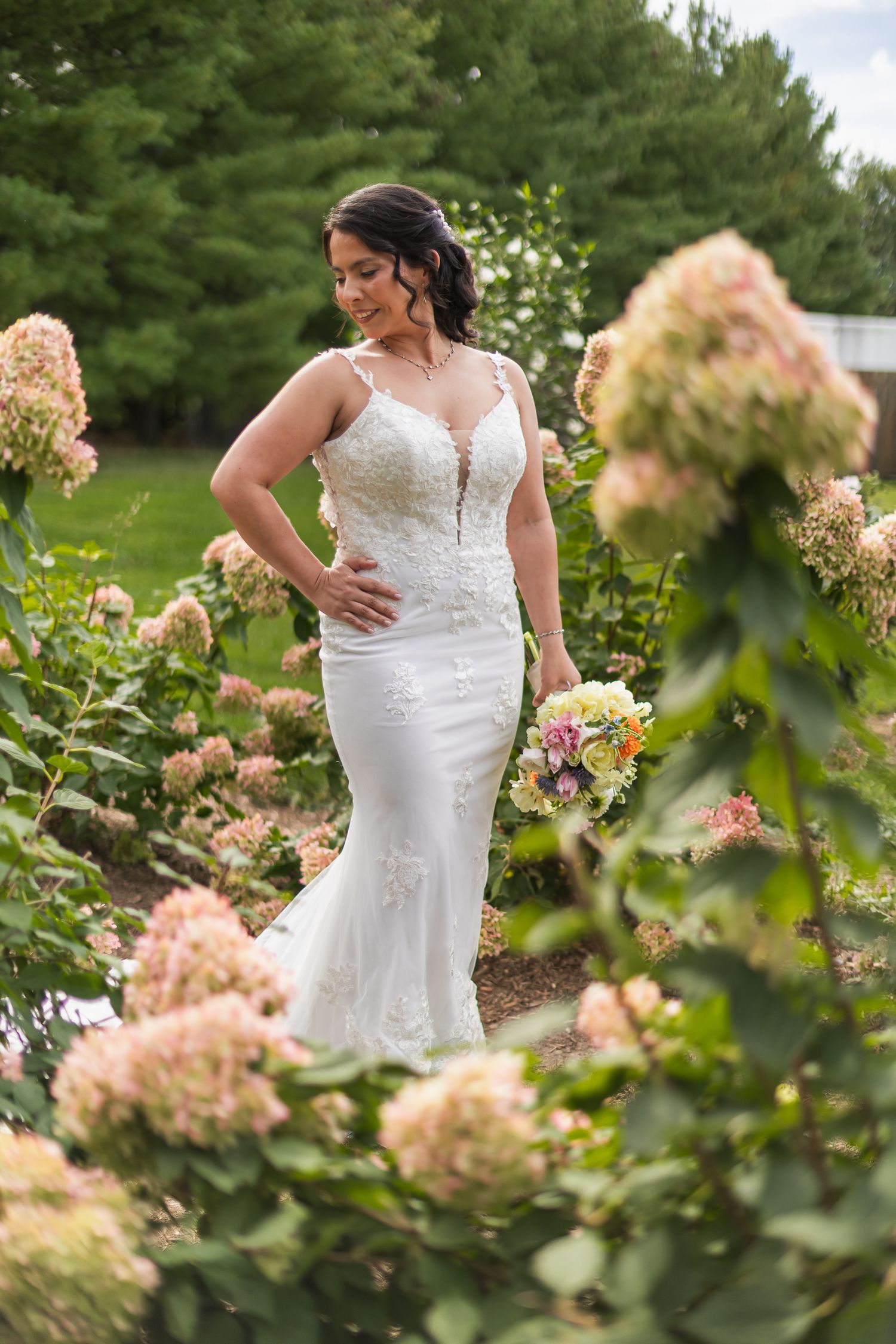 Bride in a fitted lace wedding gown posing elegantly among blooming pink hydrangeas in a garden.