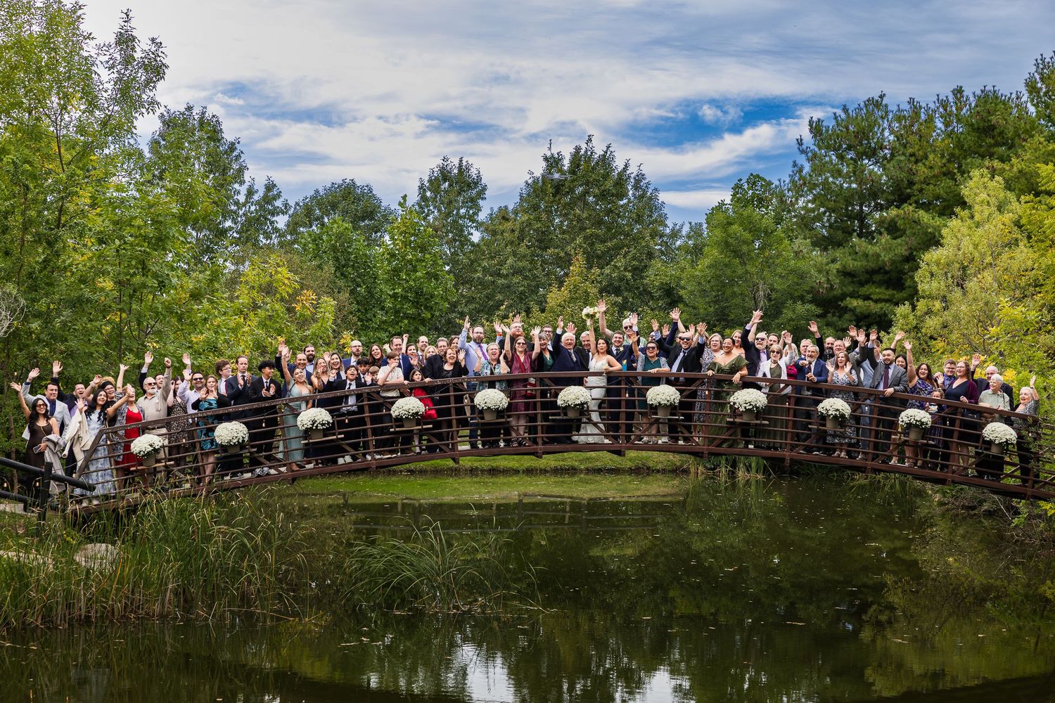 Large wedding party group photo on a bridge over a pond surrounded by lush green trees.