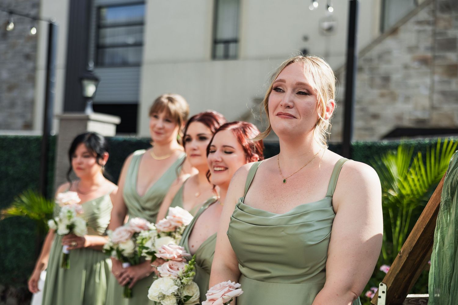 Bridesmaids in sage green dresses holding bouquets at an outdoor wedding ceremony.
