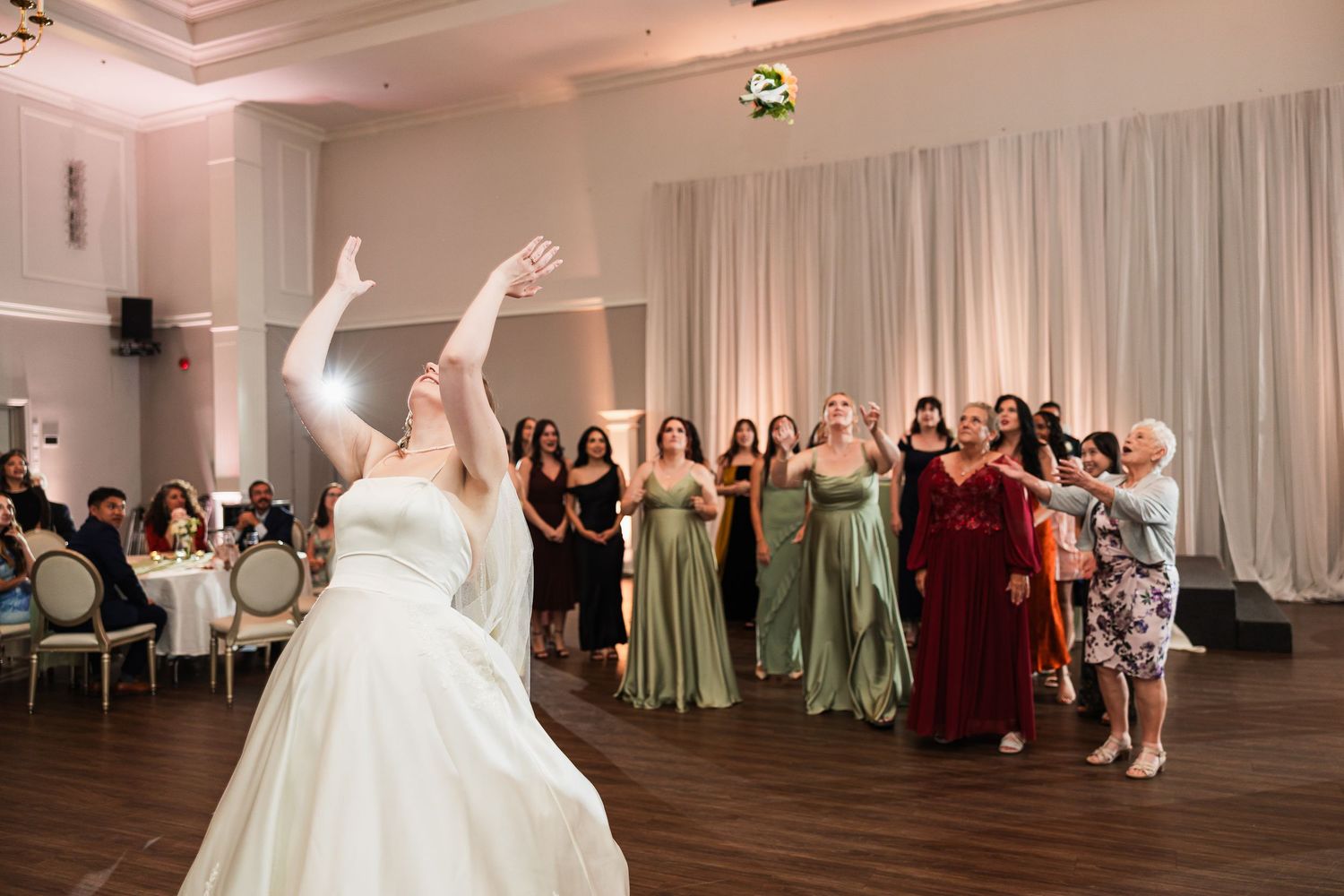 Bride tossing bouquet to group of women in green and red dresses at elegant wedding reception.