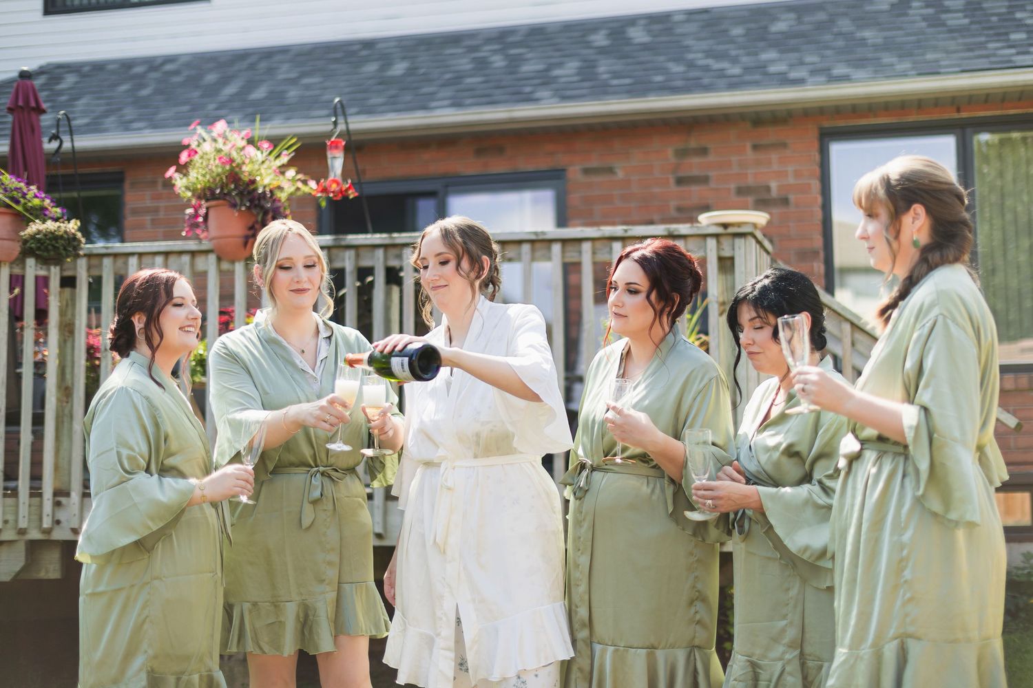 Bride pouring champagne for bridesmaids in sage green robes on a sunny day.