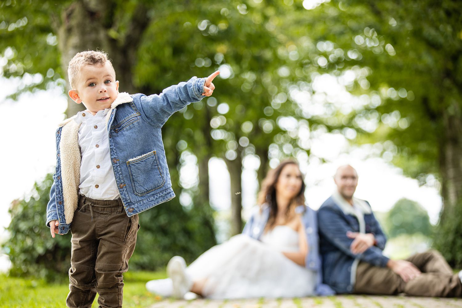 Child in blue jacket pointing while others relax on rocks in a sunny park setting.
