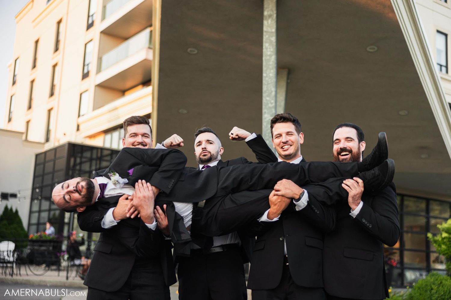 Groomsmen in black suits posing playfully outside modern building.