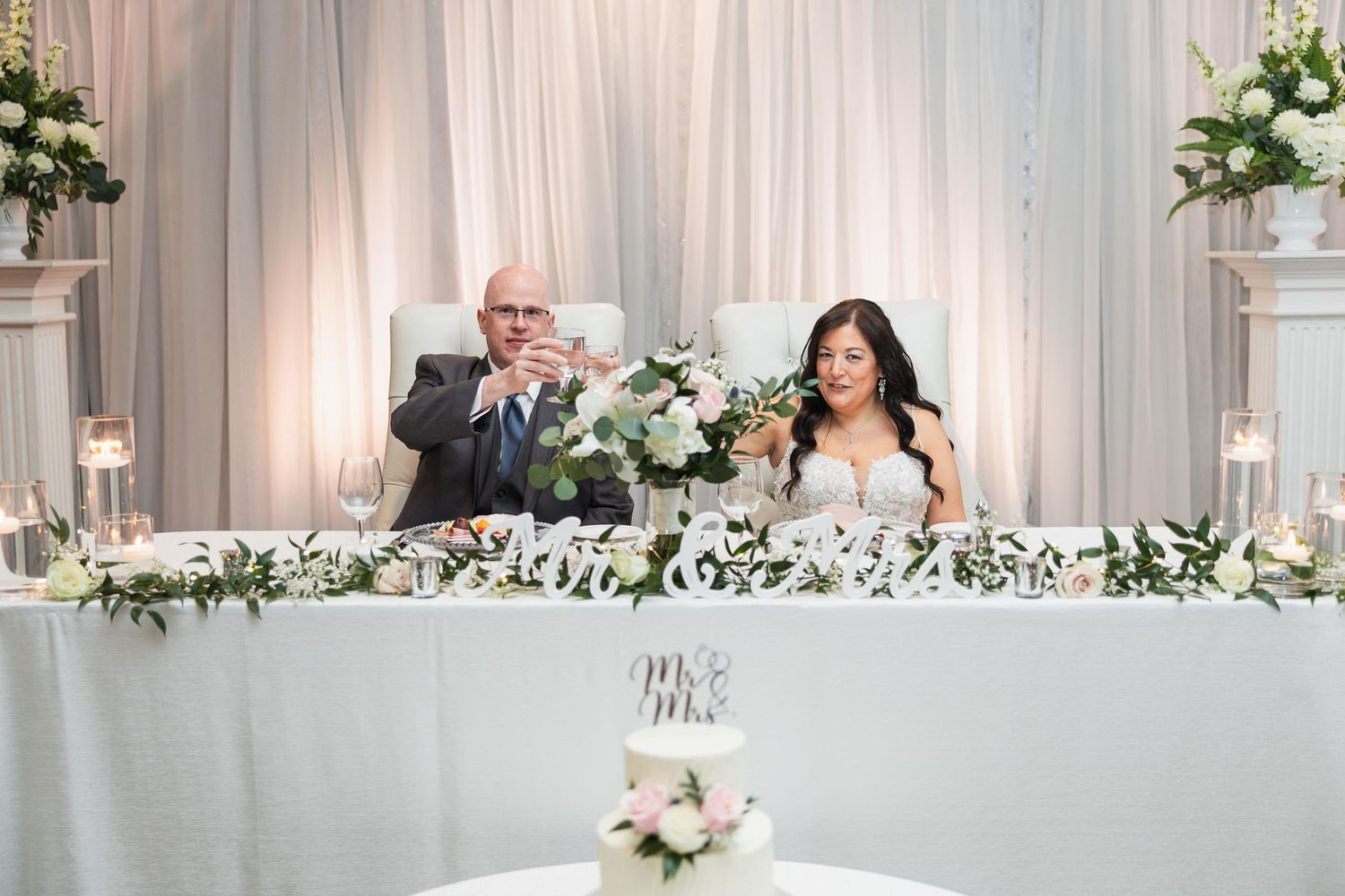 Wedding couple seated at decorated head table with white floral arrangements.