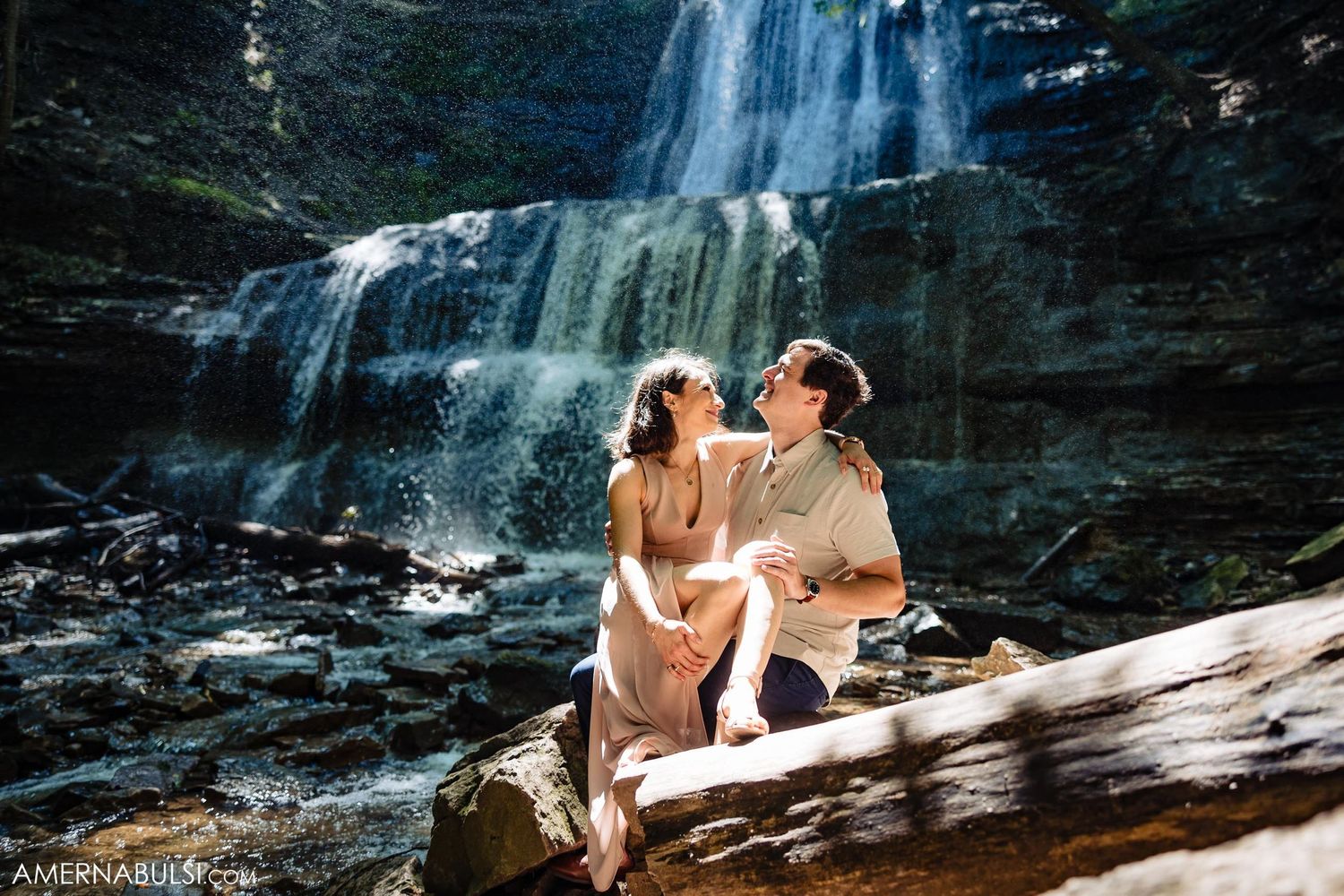 Two people share an intimate moment while sitting on a log in front of a cascading waterfall.