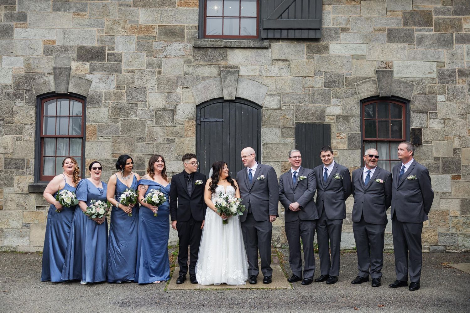 Complete wedding party poses against stone wall of historic building.