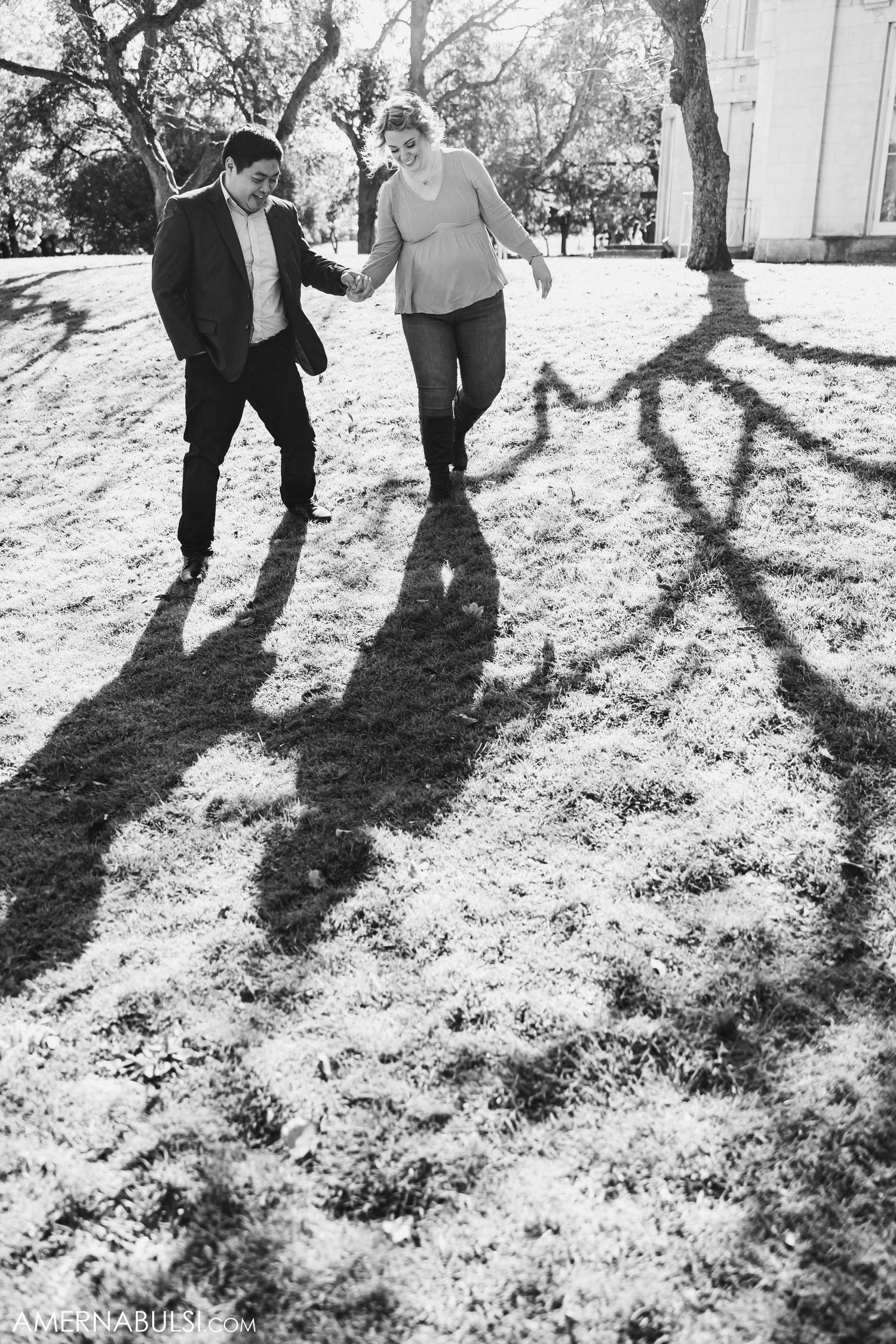 Black and white photo of couple walking playfully with dramatic tree shadows on ground.