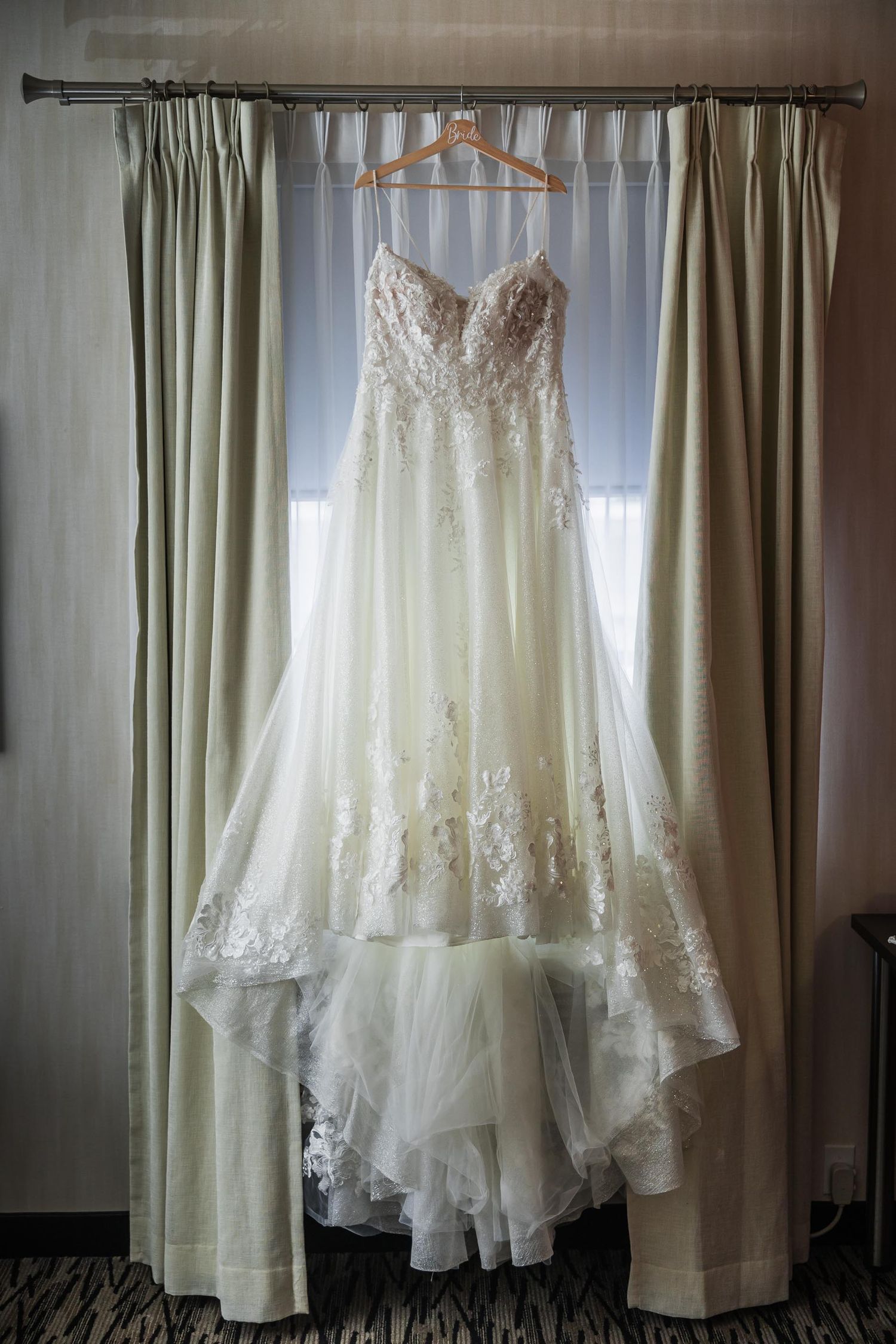 Elegant white wedding dress with lace detail hanging on wooden hanger against light curtains.