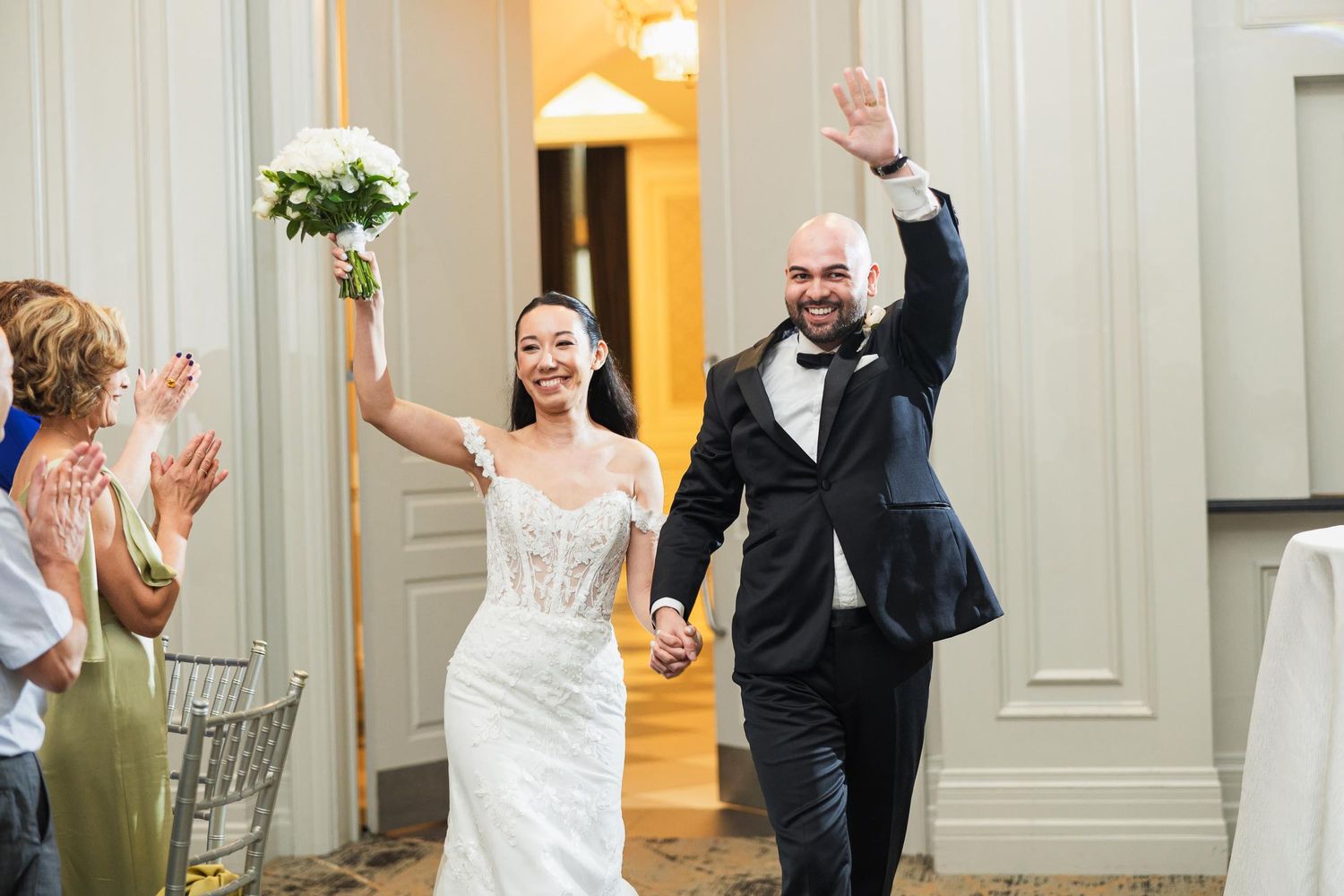 Newlyweds make their grand entrance at wedding reception while guests clap and cheer in elegant venue.
