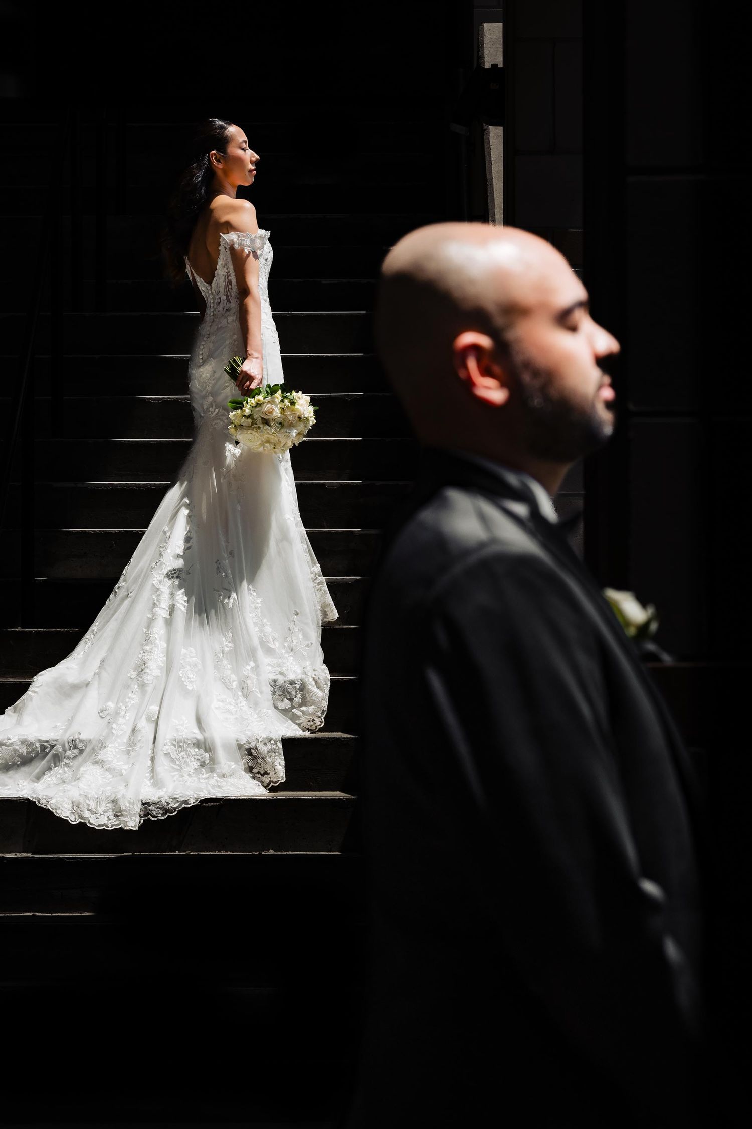 A dramatic wedding portrait on dark stairs with contrasting lighting between bride in white and groom in black.