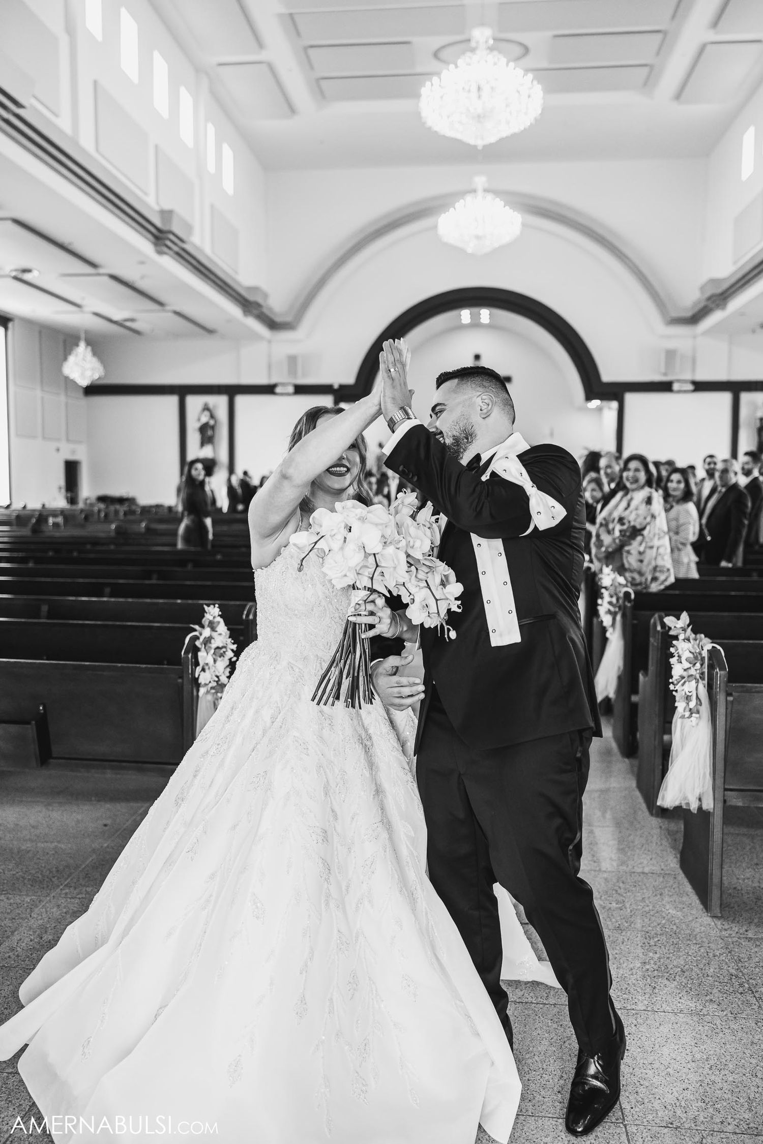 Joyful newlyweds dance down the church aisle under crystal chandeliers as guests look on.