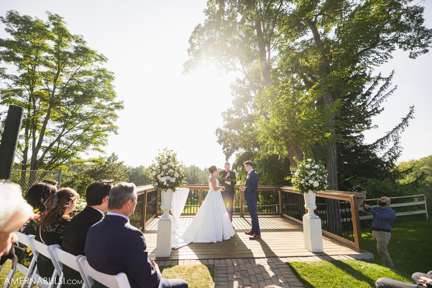 Sunset wedding ceremony on wooden deck with floral arrangements and white chairs.