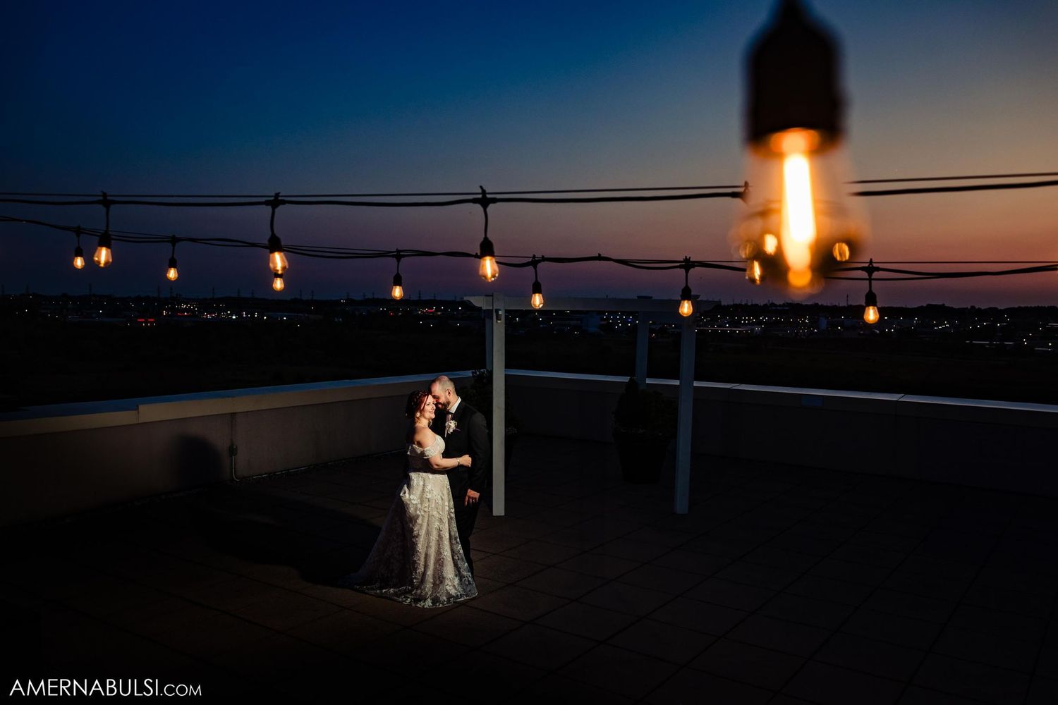 Couple embracing on rooftop terrace at dusk with string lights and lighthouse in background.