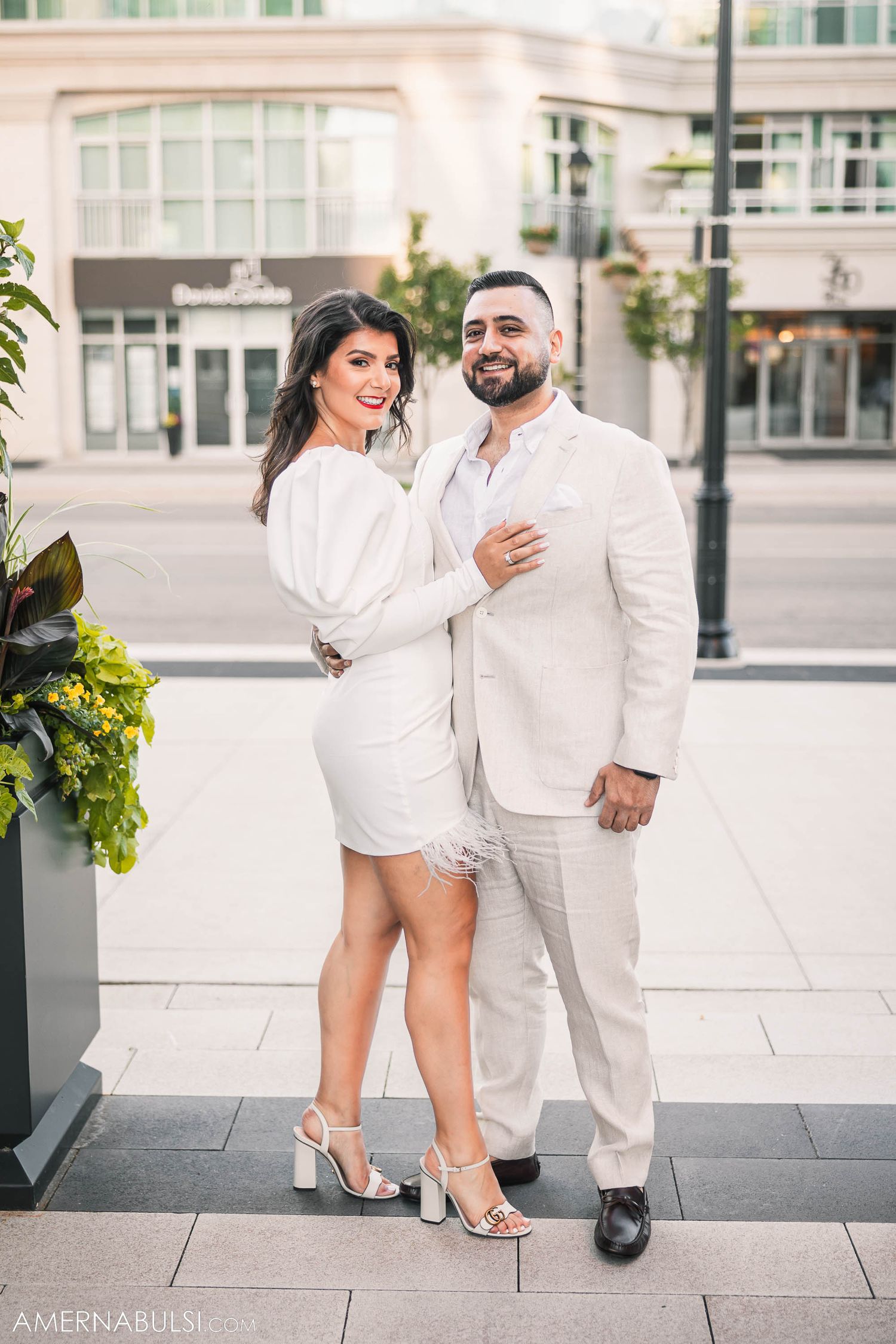 Elegant couple in all-white outfits pose together on modern urban sidewalk with planters.