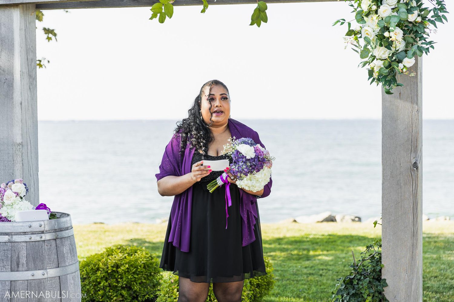 Person in purple dress holding bouquet under white floral-decorated pergola with ocean view.