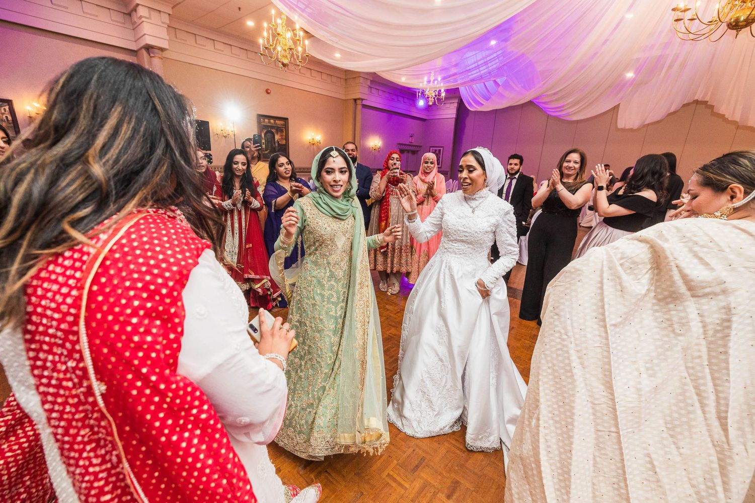 Wedding celebration with guests in colorful traditional attire dancing under purple lighting.