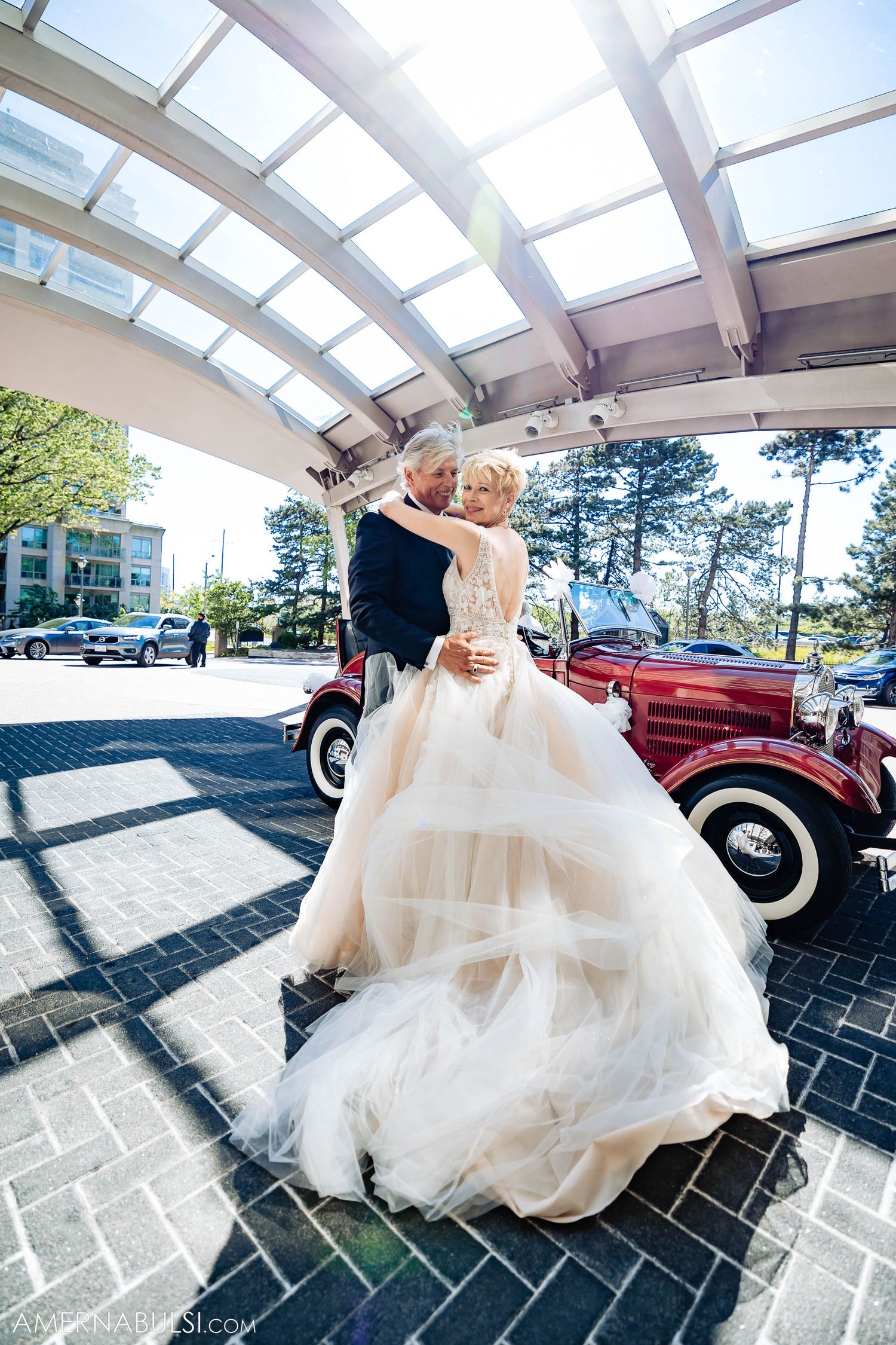 A formal couple dancing next to a vintage red car under a modern glass ceiling structure.