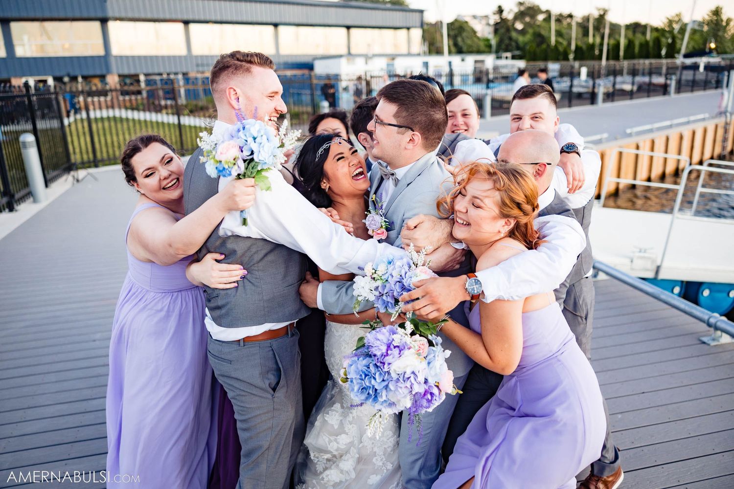Wedding party celebrates with laughter and hugs on an outdoor deck with purple bridesmaids dresses and blue bouquets.