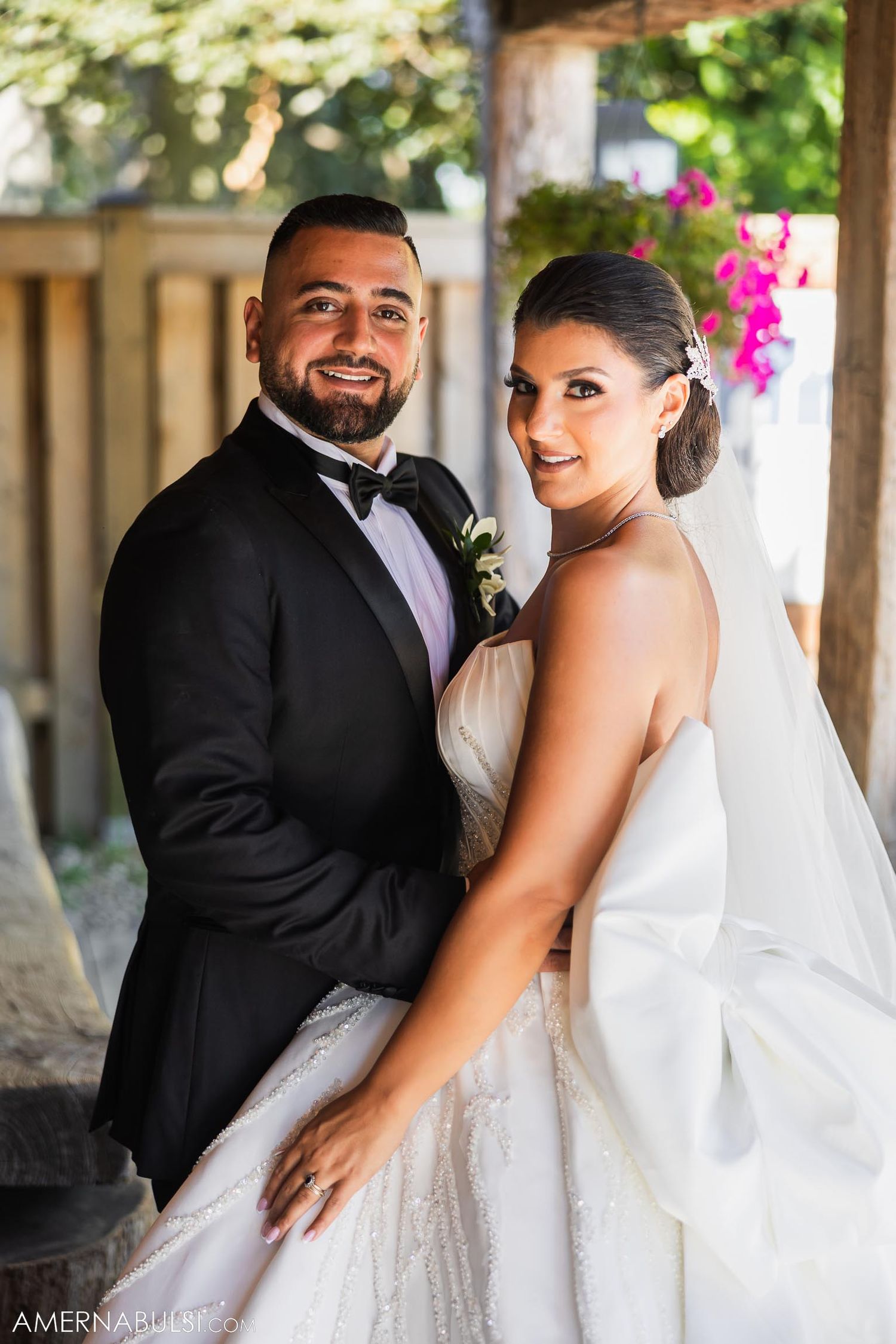 A couple poses together under a covered porch decorated with pink flowers on their wedding day.
