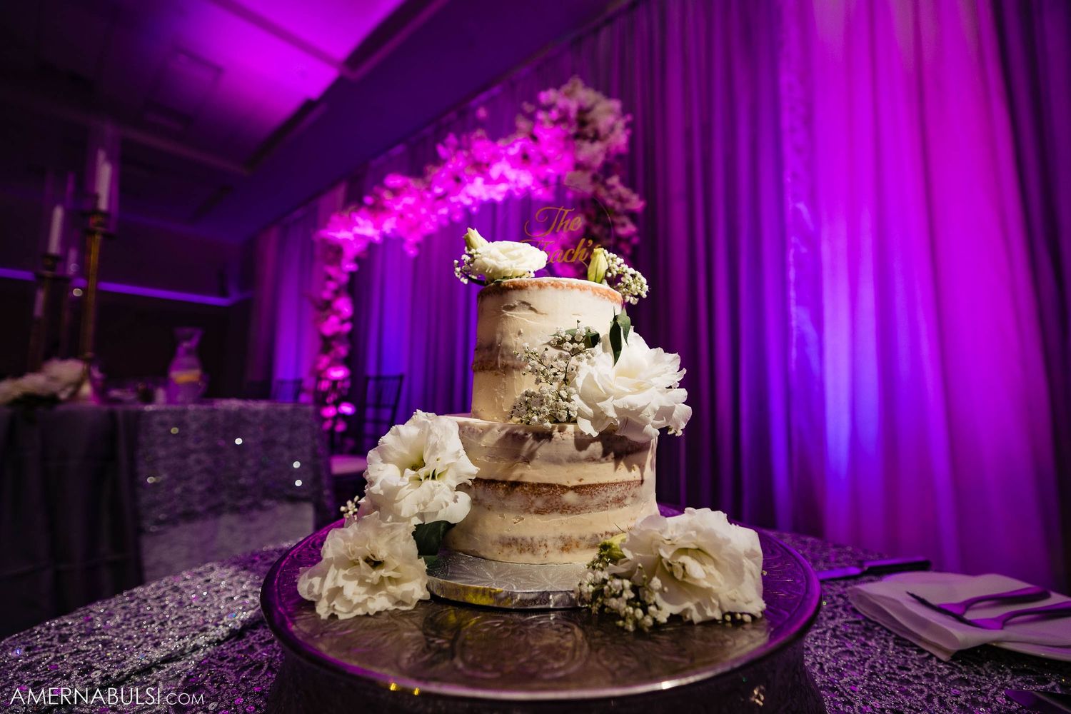Elegant wedding cake with white flowers displayed under dramatic purple lighting at reception.