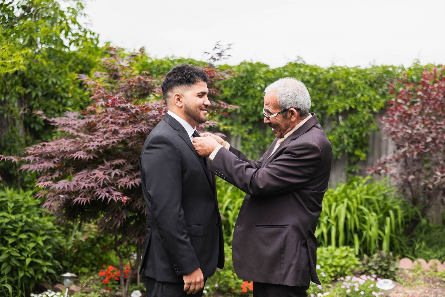 An emotional moment as an older man adjusts a younger man's formal attire in a garden setting.