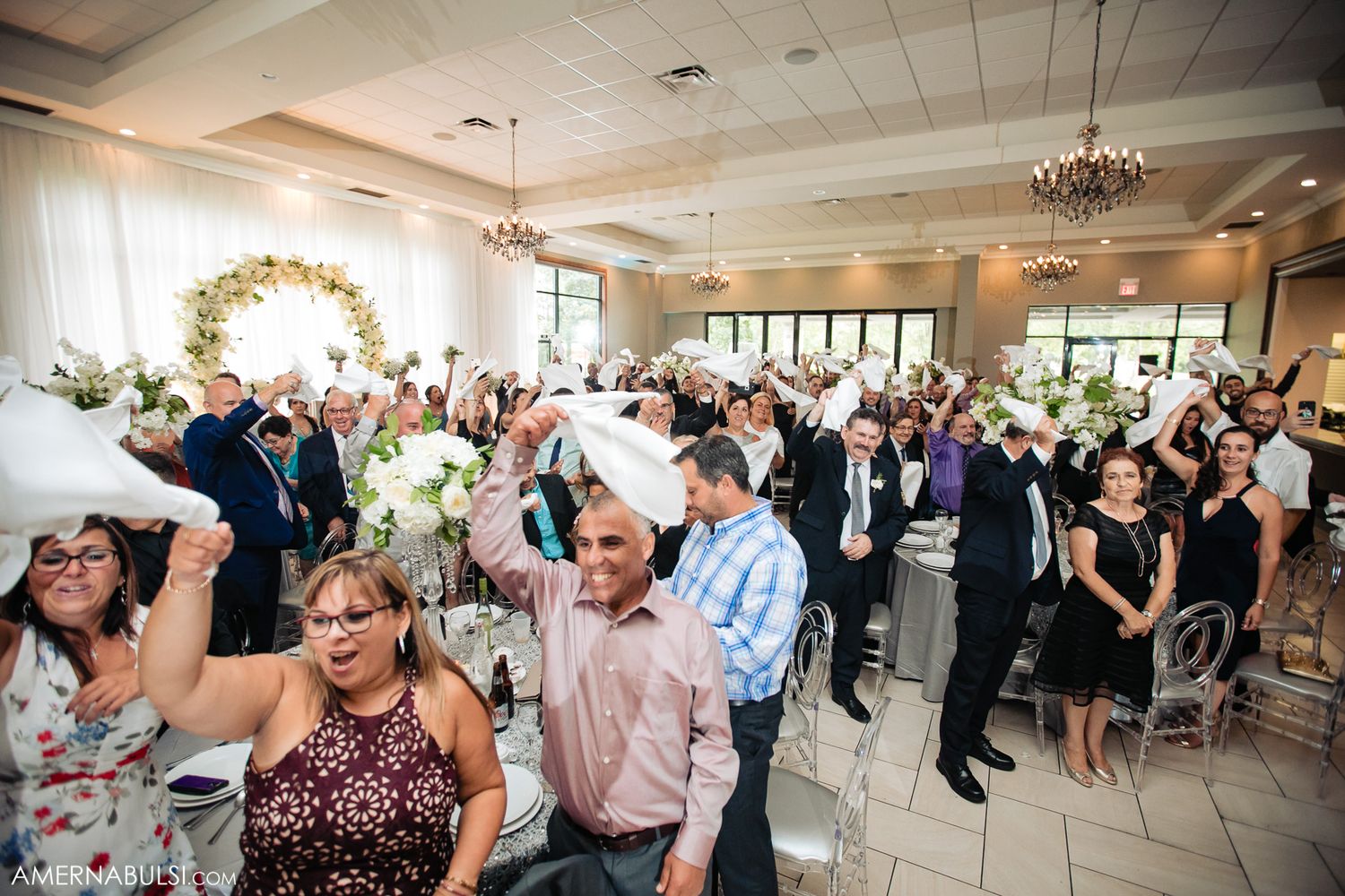 Wedding guests dancing and celebrating in a beautifully decorated reception hall with floral arrangements.