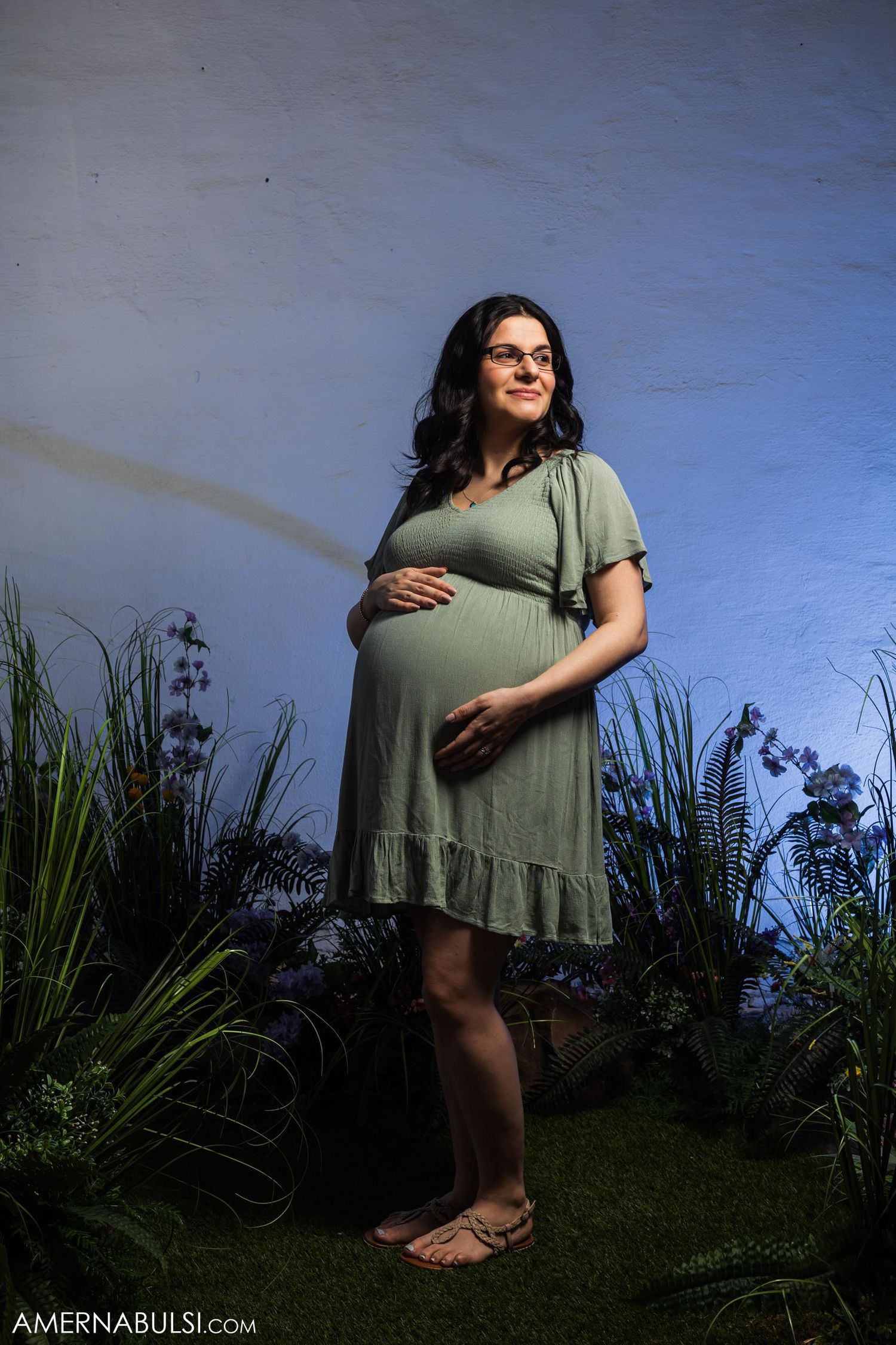 Maternity portrait with woman in sage green dress standing among garden plants at dusk.