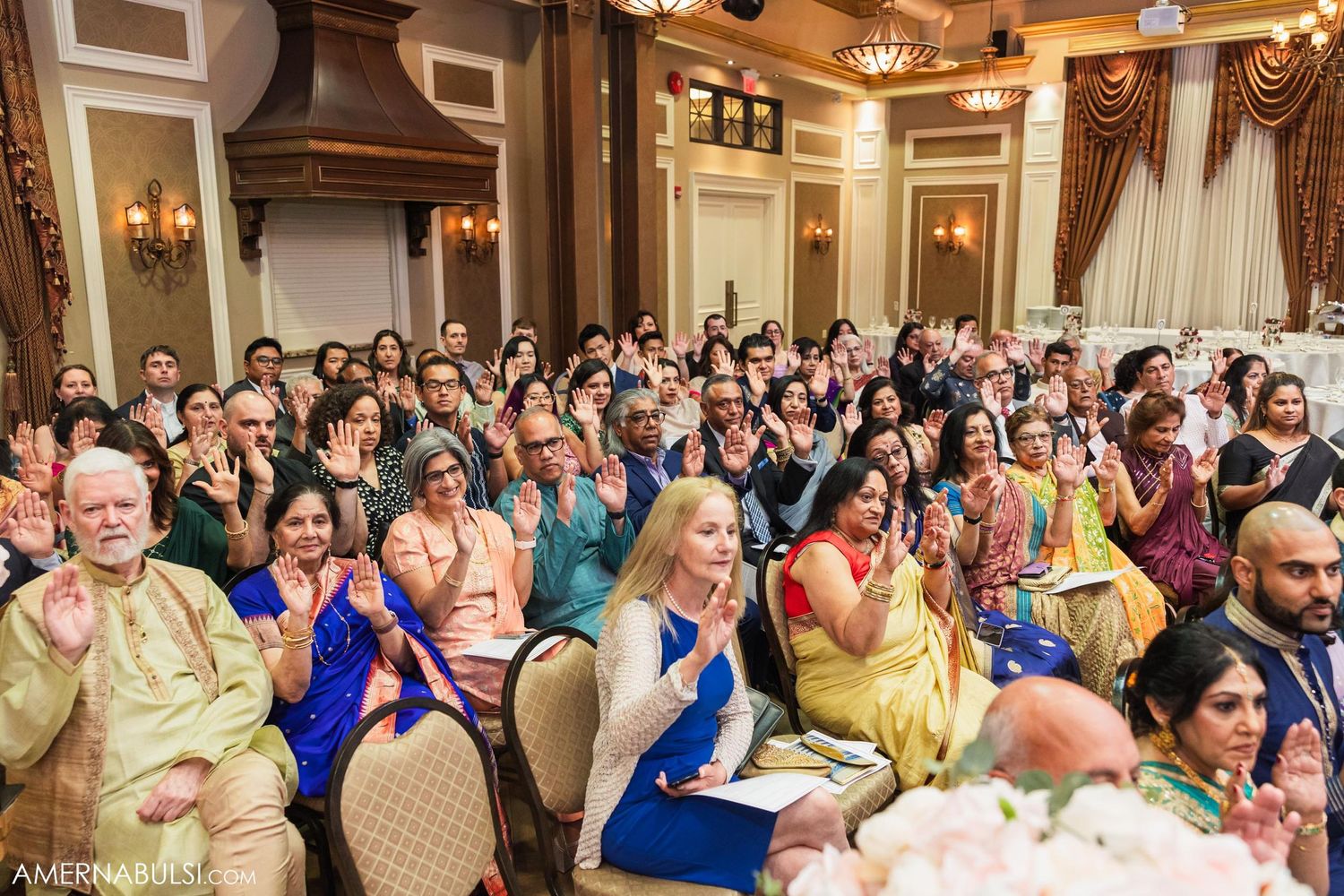 A large crowd of wedding guests in colorful traditional attire watch the ceremony from their seats.