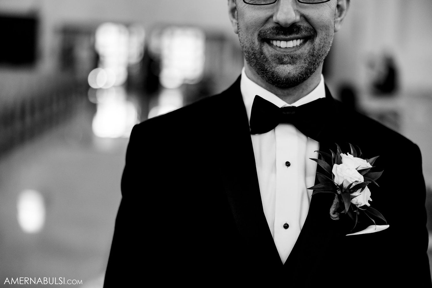 A close-up shot of a groom wearing a black tuxedo and bowtie with a boutonniere at a formal wedding.