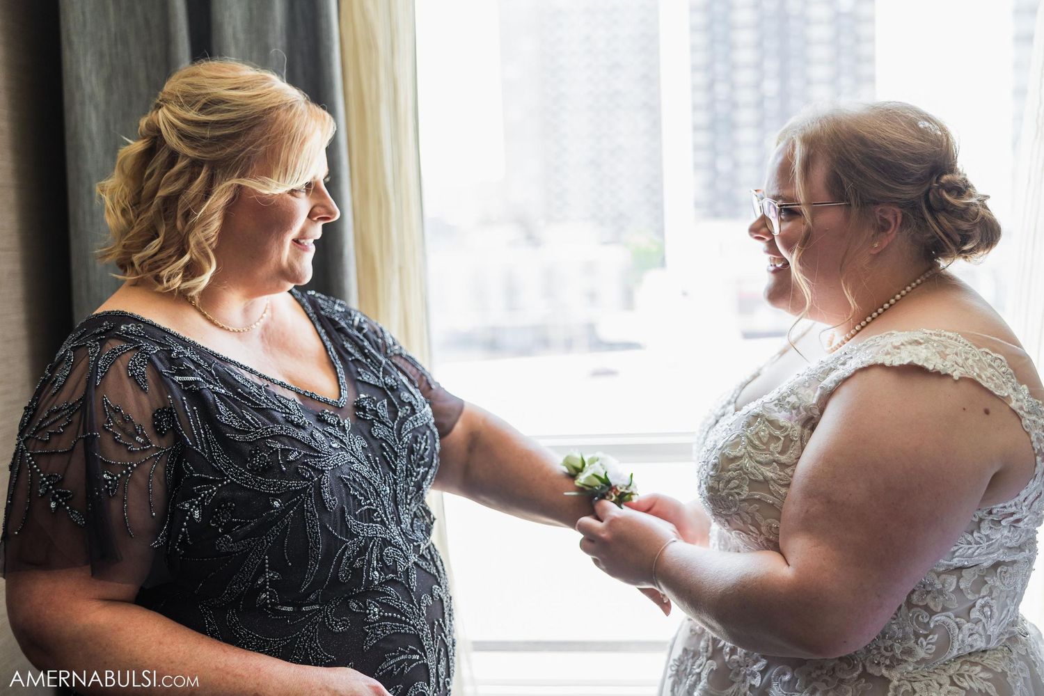 Two women share intimate moment while getting ready in hotel room with city view.