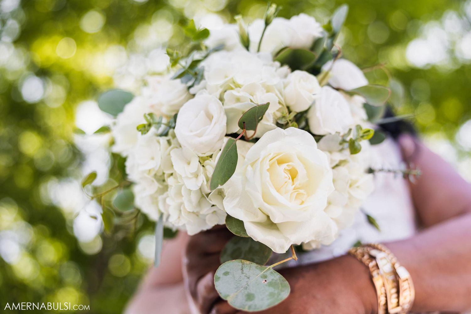 Elegant white rose bouquet held against a bokeh green background with wedding rings visible.