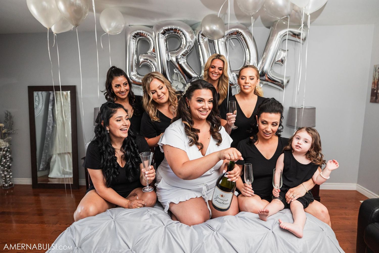 Group in black robes poses on bed under silver BRIDE balloon decorations.