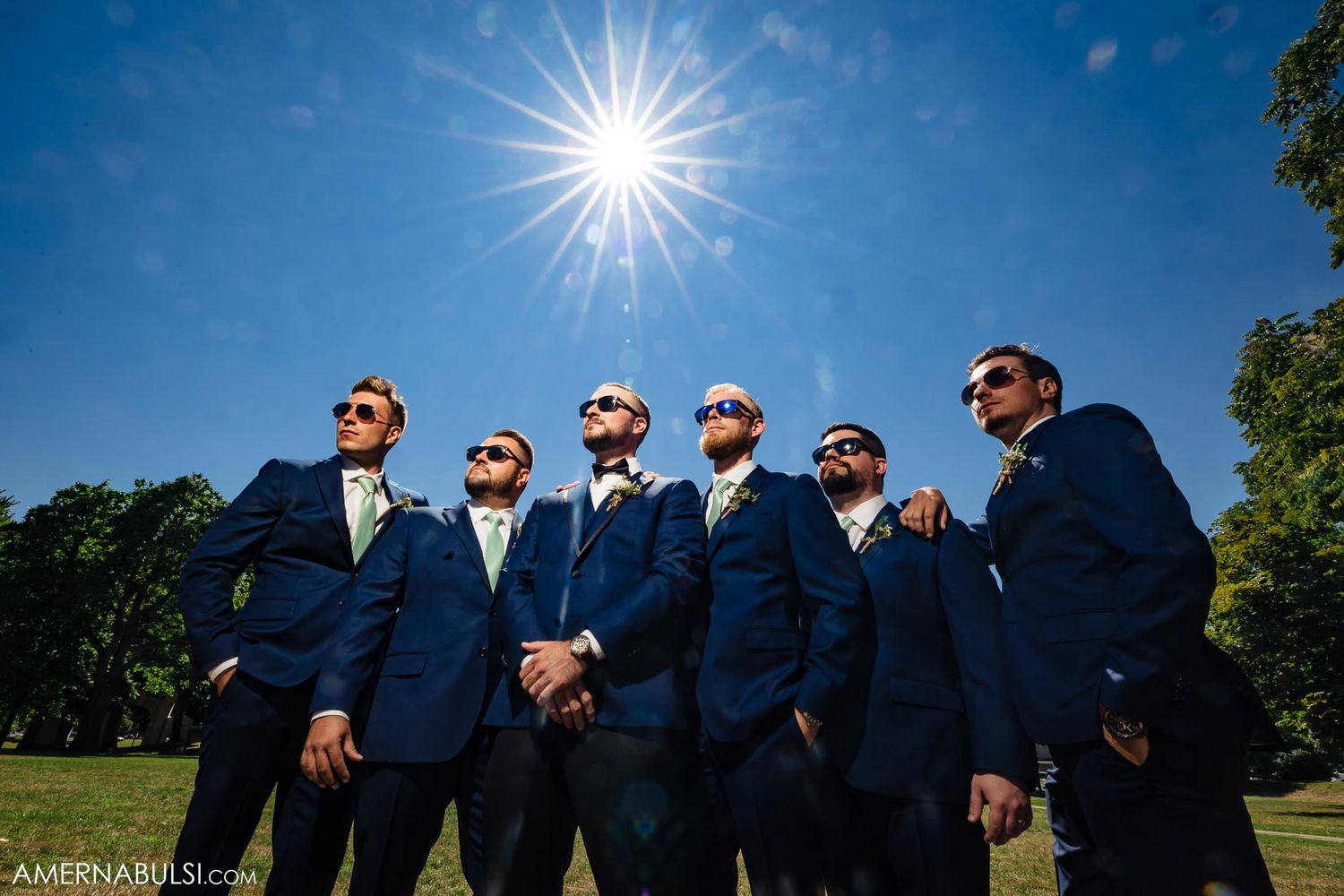 Groomsmen in matching navy suits posing against bright sunburst on a sunny day.