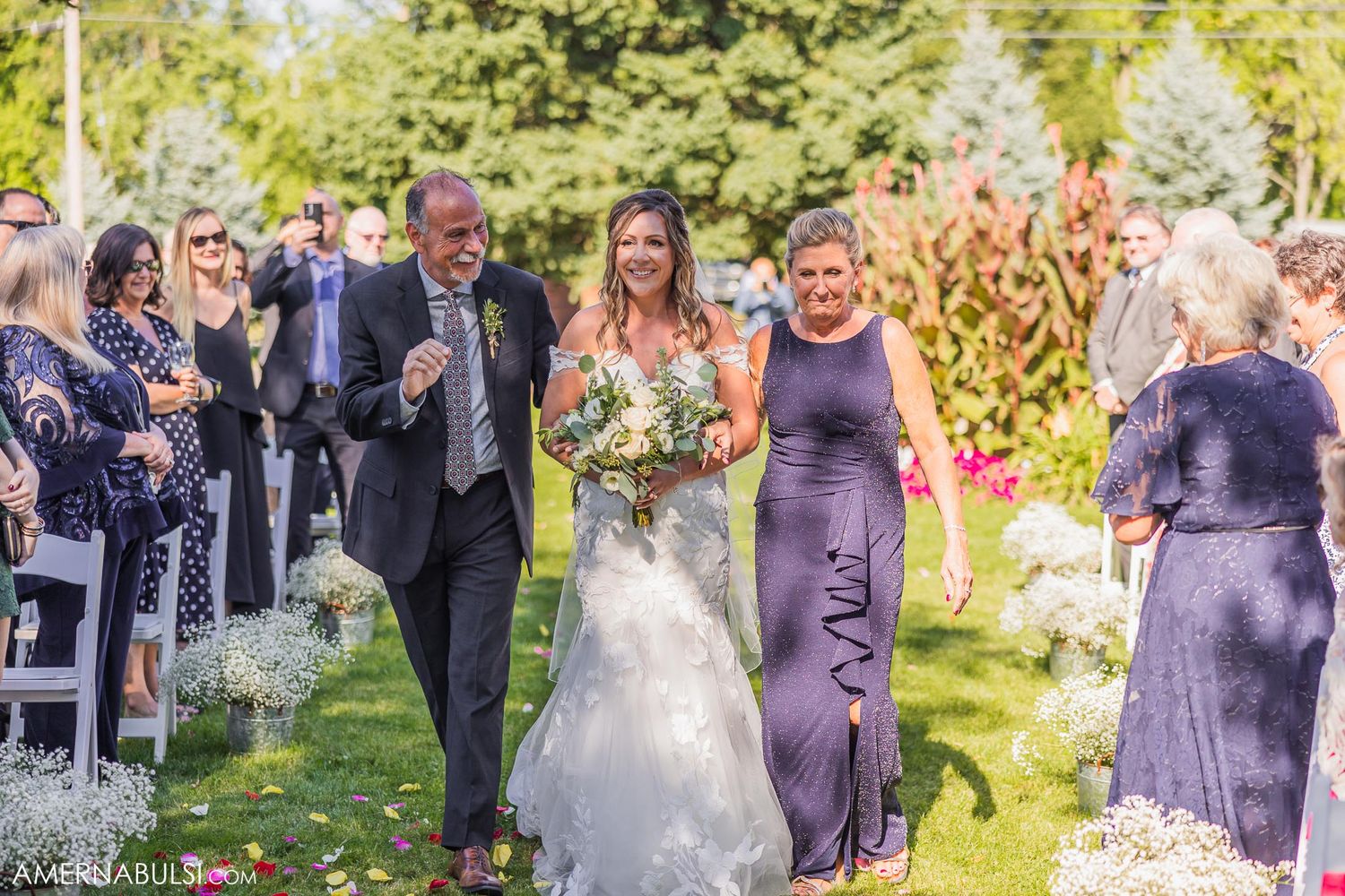 Wedding procession walking down outdoor aisle with guests in formal attire.