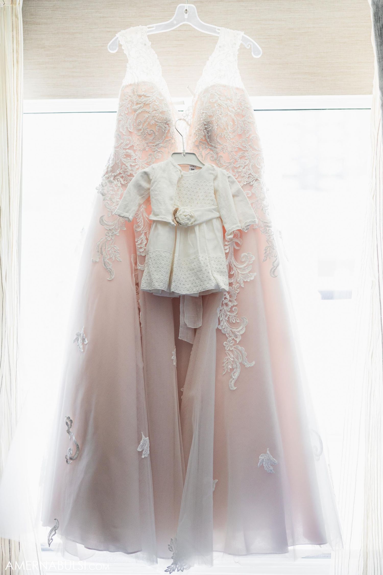 Blush pink wedding gown with lace detail and small white outfit hanging in window.
