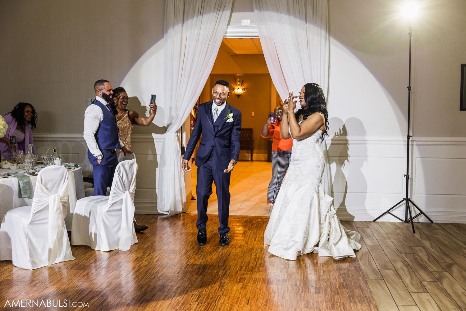 Wedding party entrance through white draped archway with guests watching from decorated reception hall.