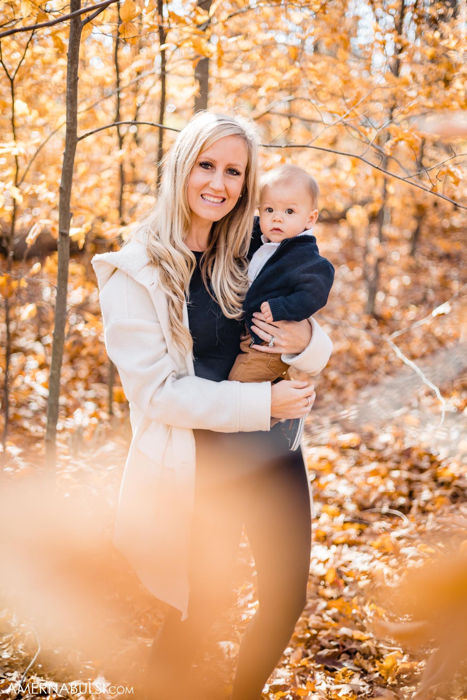 Parent holds smiling baby against backdrop of golden autumn trees.